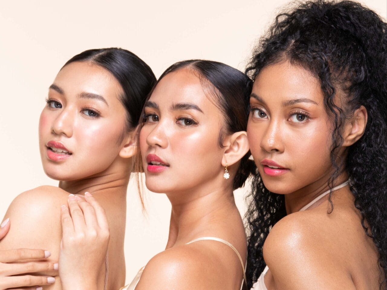Three women with glowing skin pose together, showcasing natural beauty with varied hairstyles against a neutral beige background.