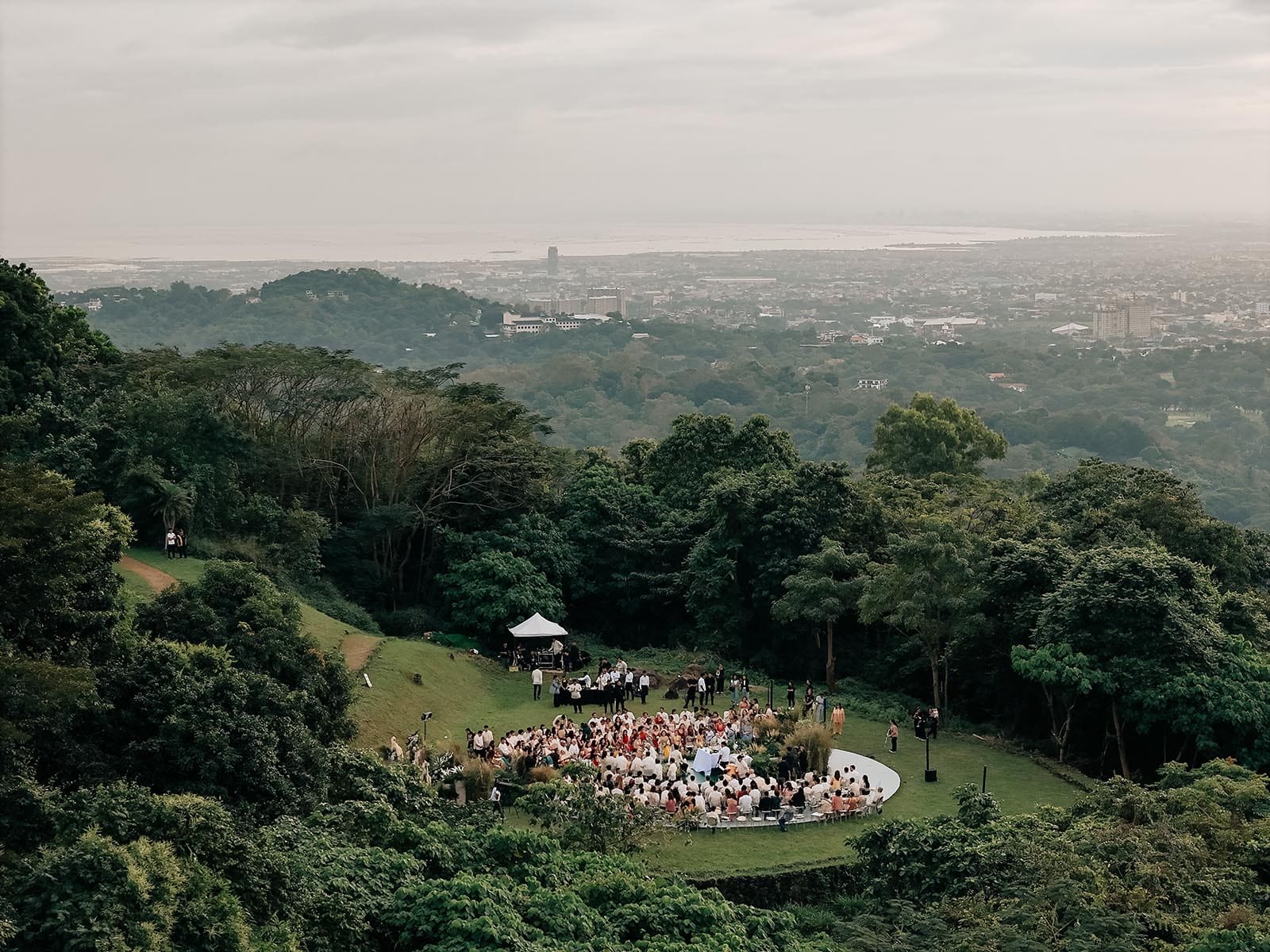 The image shows an overhead photo of Kiana Valenciano and Sandro Tolentino's wedding, set on a lush hilltop with a panoramic view of the city. Guests are seated in a circular arrangement on a grassy lawn, surrounded by greenery.