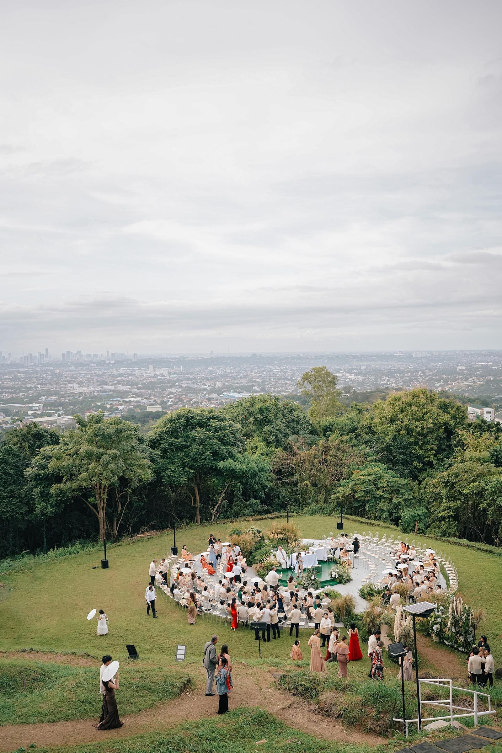 The image shows an overhead photo of Kiana Valenciano and Sandro Tolentino's wedding, set on a lush hilltop with a panoramic view of the city. Guests are seated in a circular arrangement on a grassy lawn, surrounded by greenery.