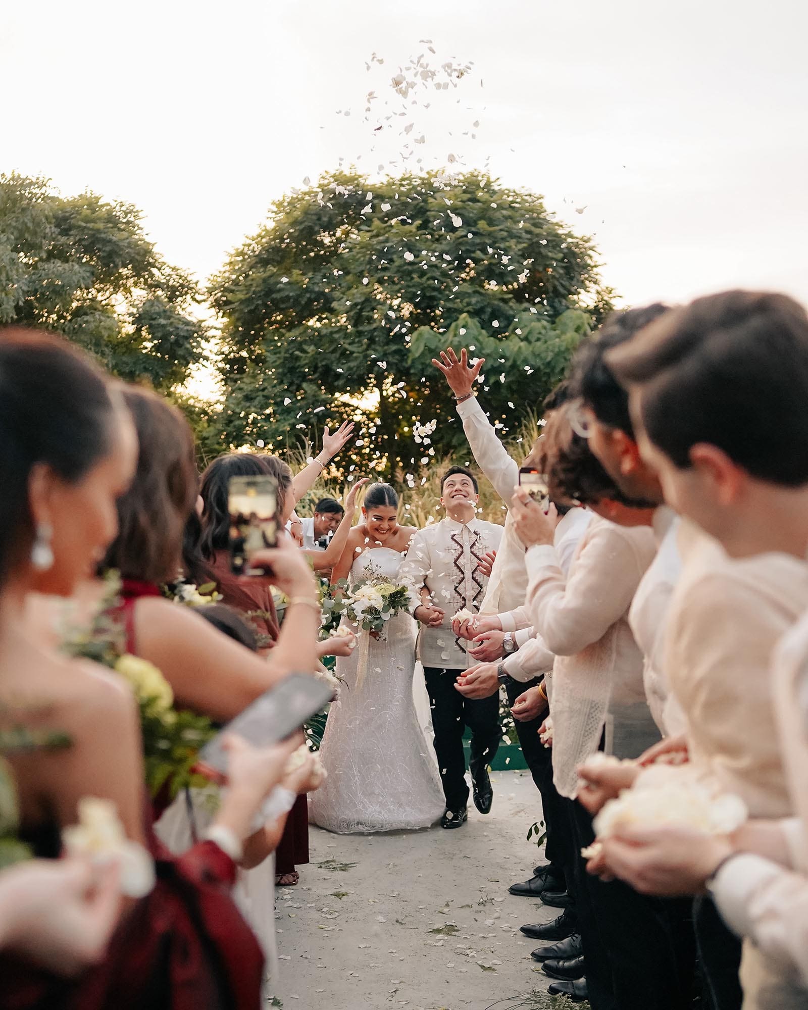 The image captures Kiana Valenciano and Sandro Tolentino walking hand-in-hand as newlyweds through a joyous aisle lined with friends and family. Guests are throwing flower petals into the air, celebrating the couple's union. Kiana wears a radiant white wedding gown, holding a bouquet, while Sandro dons a traditional barong.