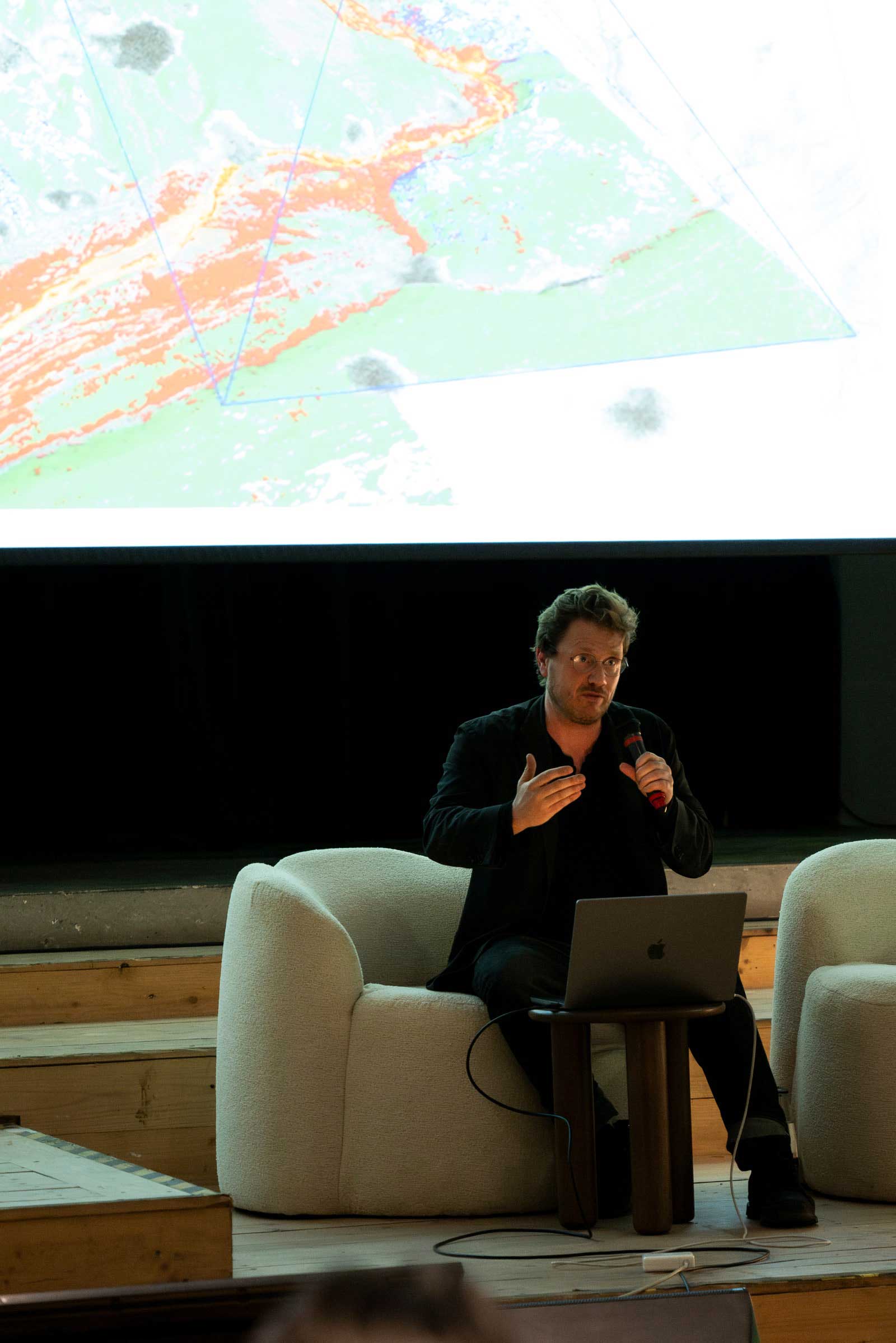 A man seated in front of his laptop holding a mic speaking to an audience.