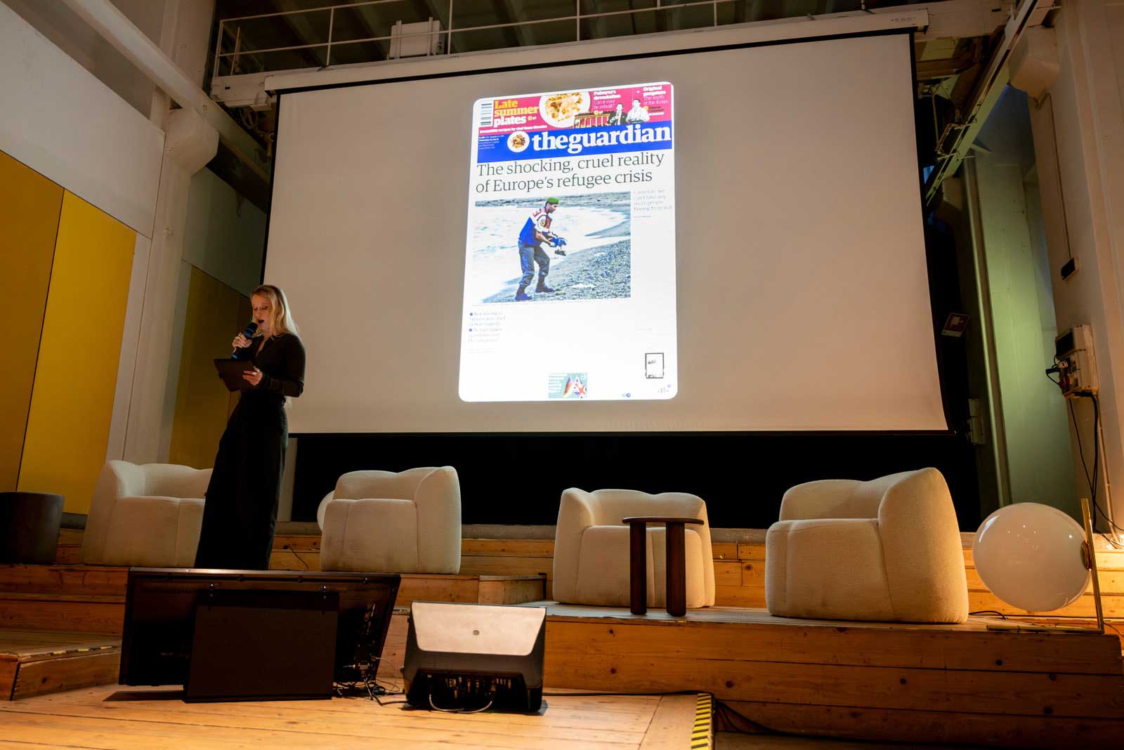 A woman talking to a mic, with a newspaper article projected behind her that reads 