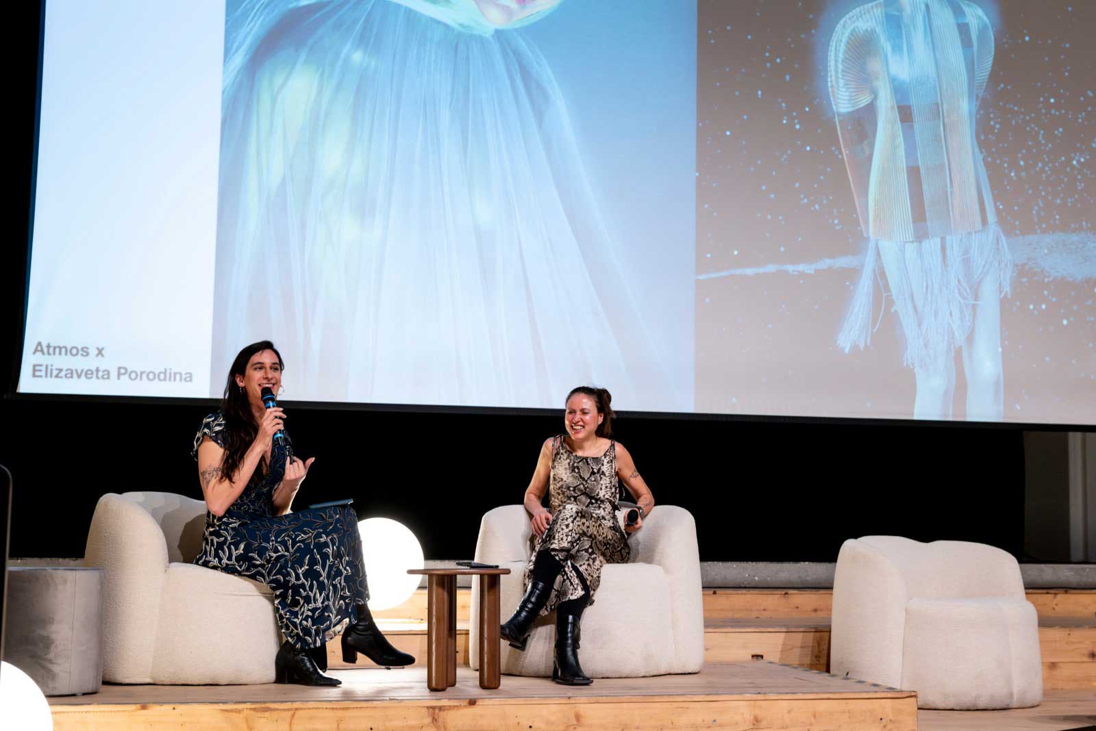 Two women seated on couches presenting to an audience.
