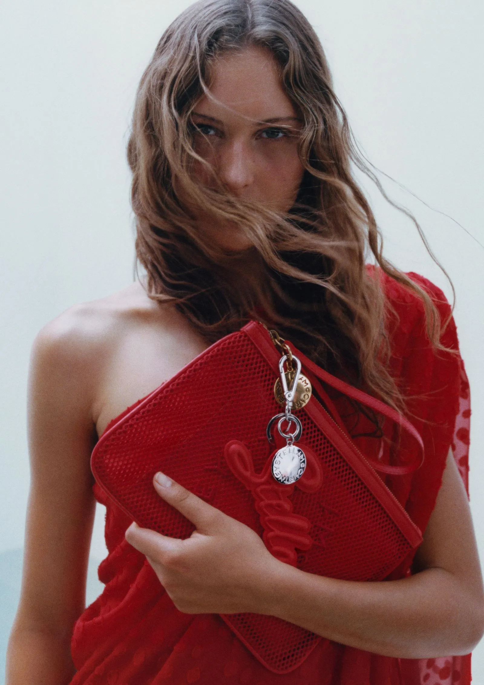Model in a red dress holding a red textured clutch with metal charms, hair blown by the wind.