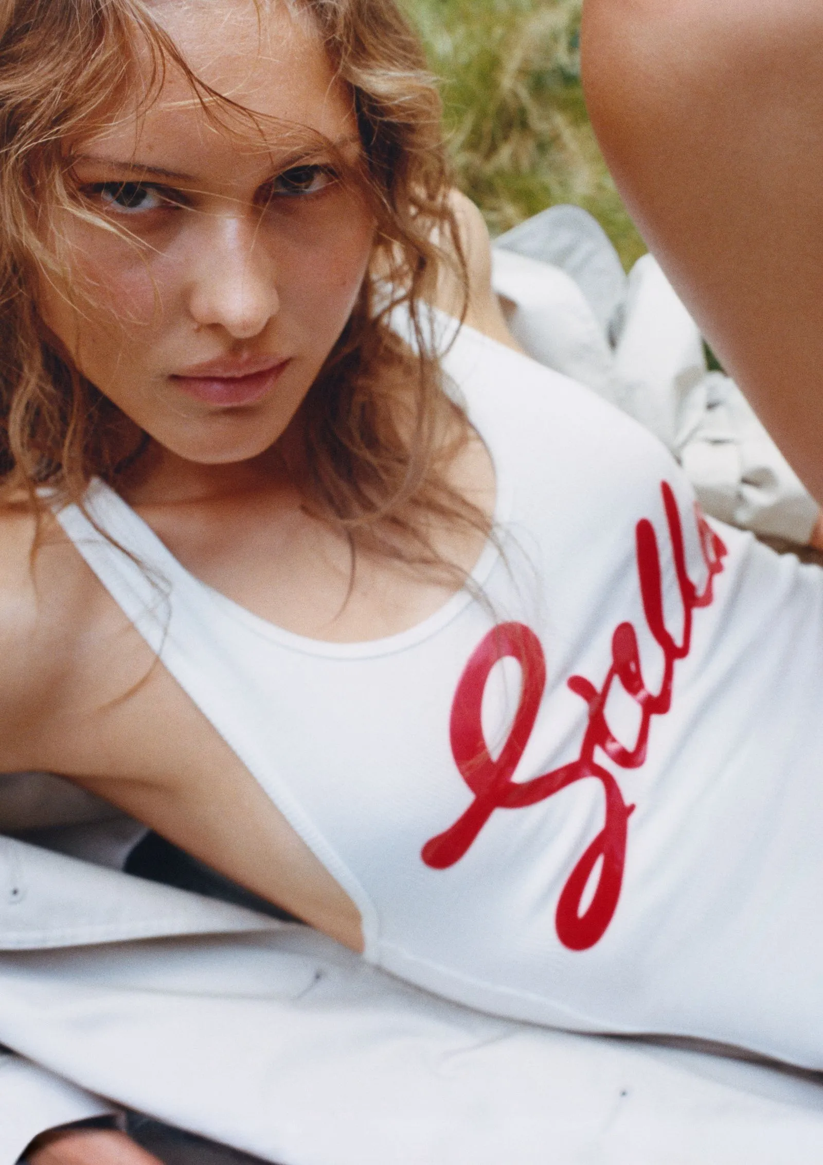 Close-up of a young woman with wavy light brown hair, wearing a white tank top with red script, outdoors.