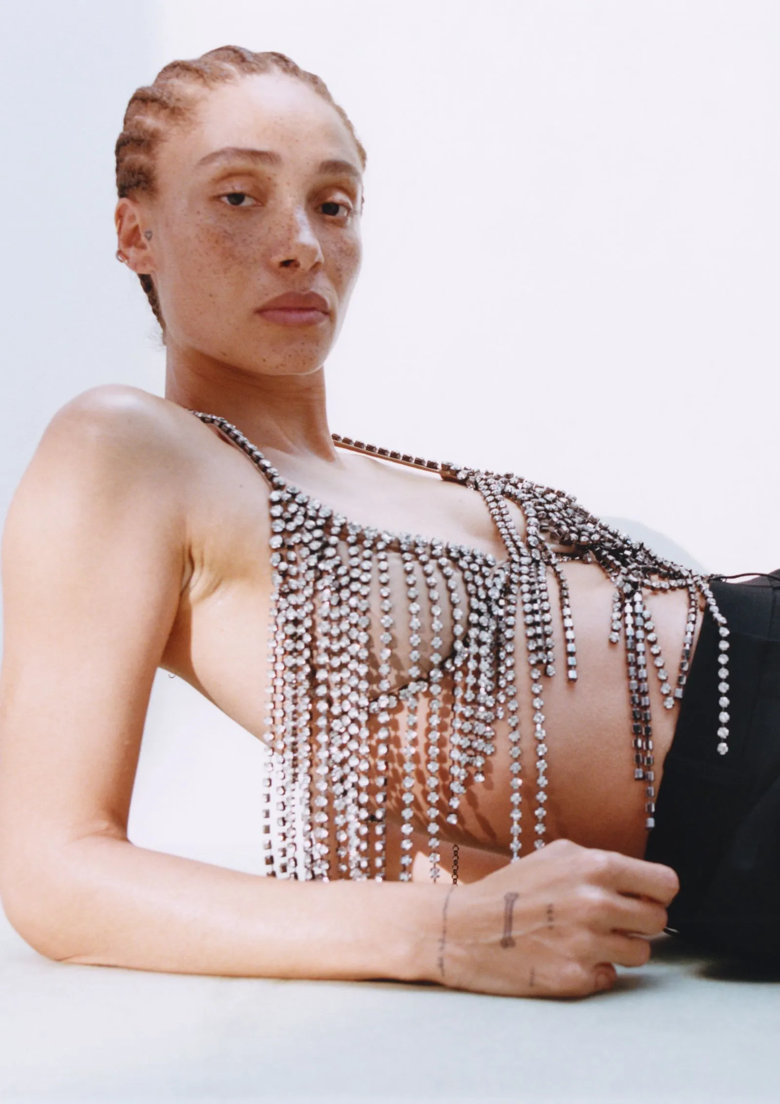 Model reclining in a sheer, crystal-beaded fringe top with chains draping over the chest.**NOTE**: body language and fashion-forward styling are the focus, set against a light background.