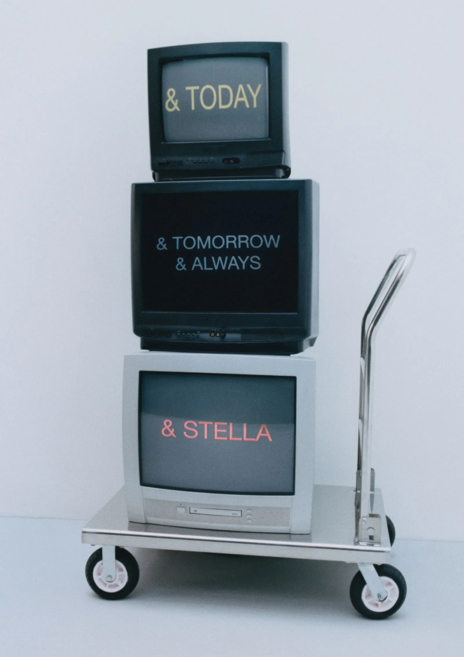 Three CRT monitors stacked on a wheeled cart displaying the phrases '& TODAY', '& TOMORROW & ALWAYS', and '& STELLA'.