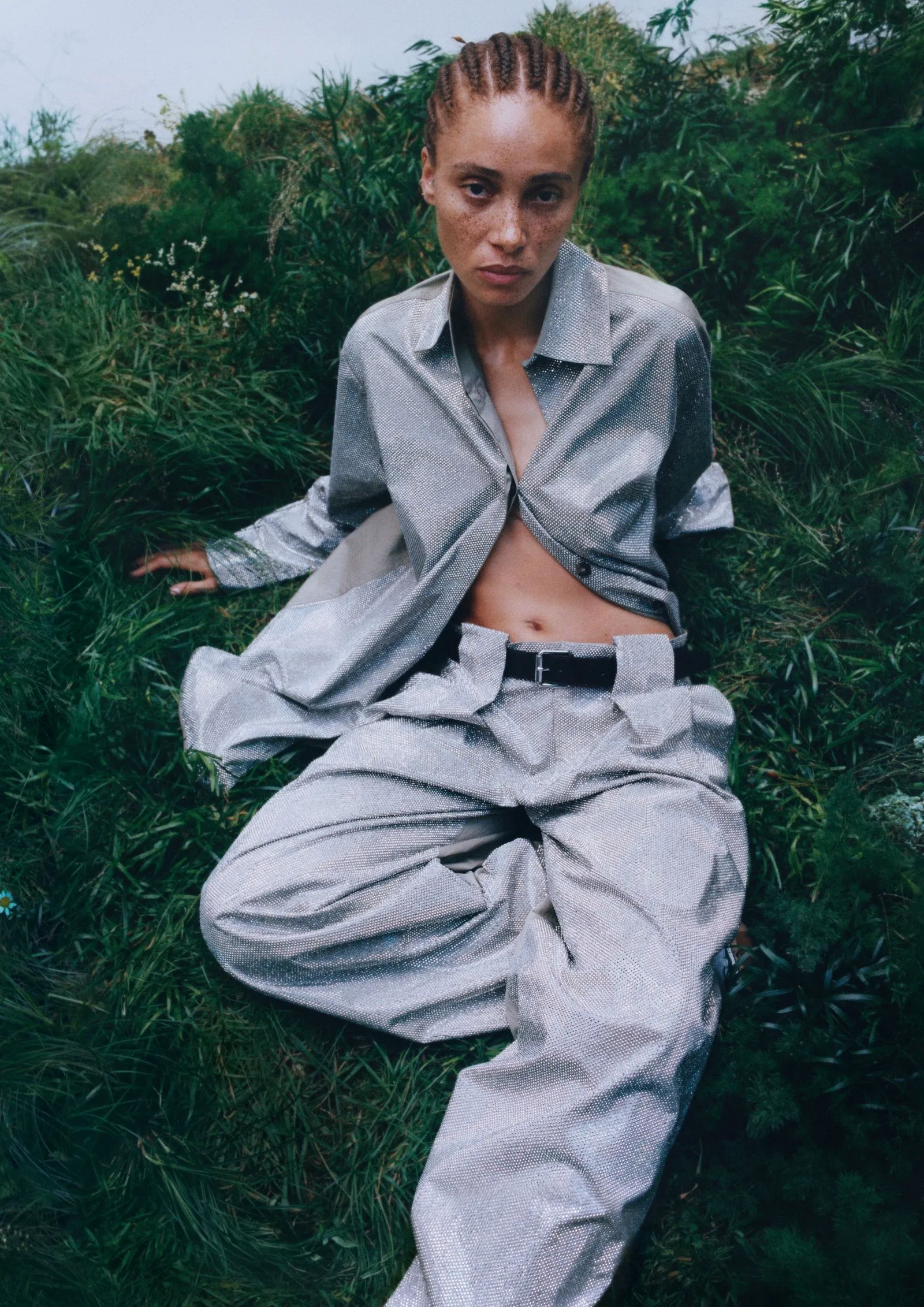 Model seated in tall grass wearing a metallic silver pantsuit with a partially unbuttoned shirt, braided hair and a focused expression.