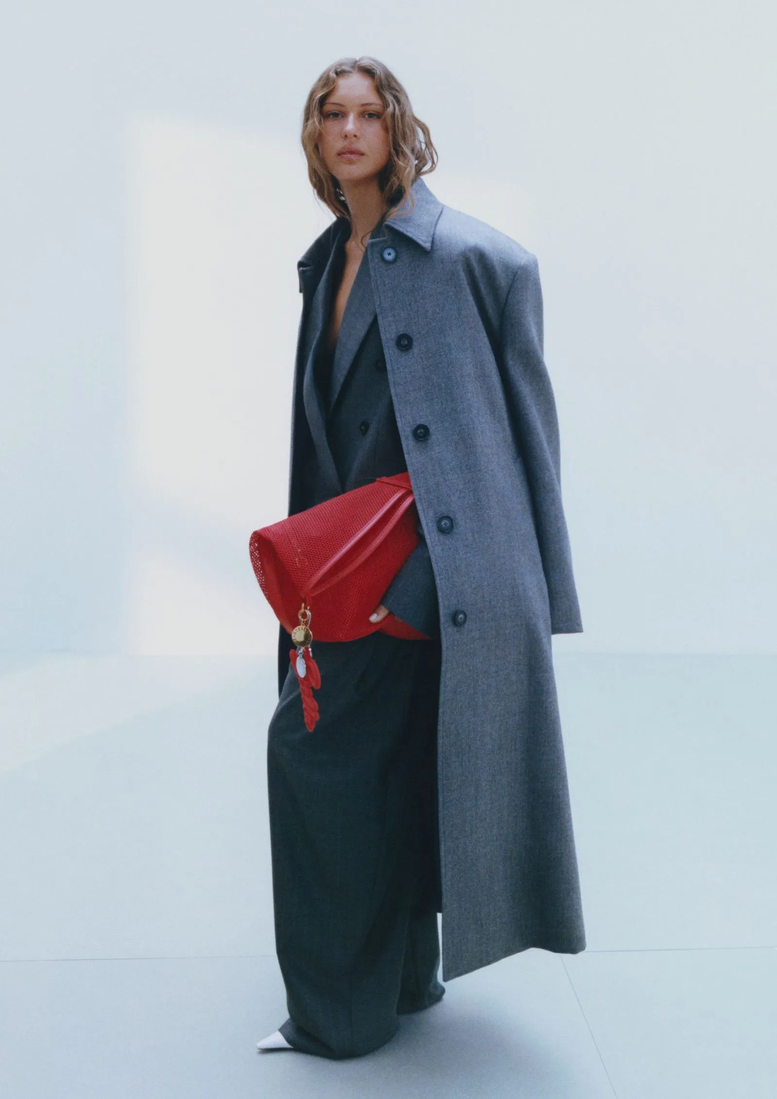 Model wearing a long gray overcoat over a dark suit, holding a bright red clutch with a gold keychain detail.