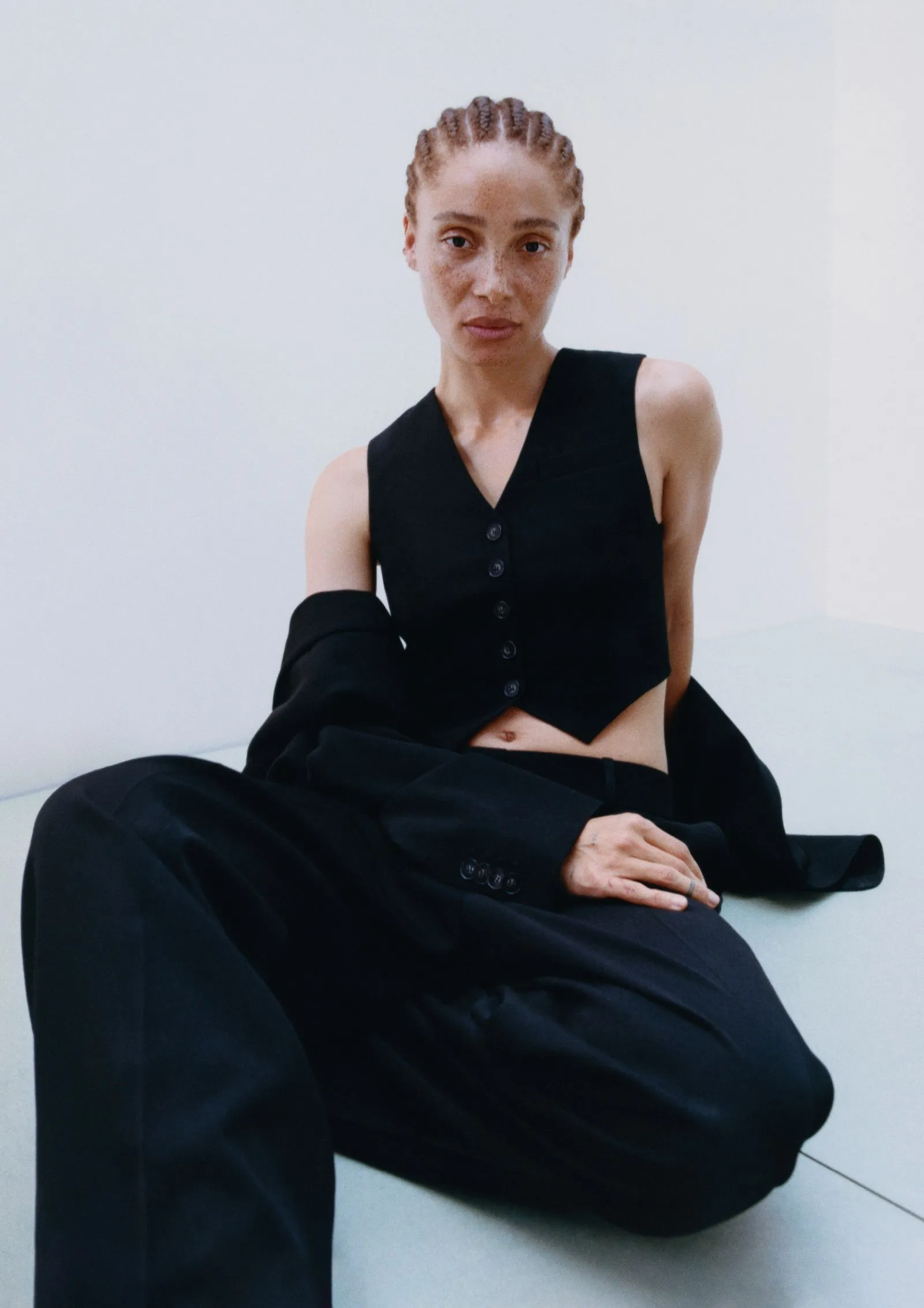 Young woman with cornrows sits on the floor in a black sleeveless vest and oversized black pants, looking at the camera against a light background.