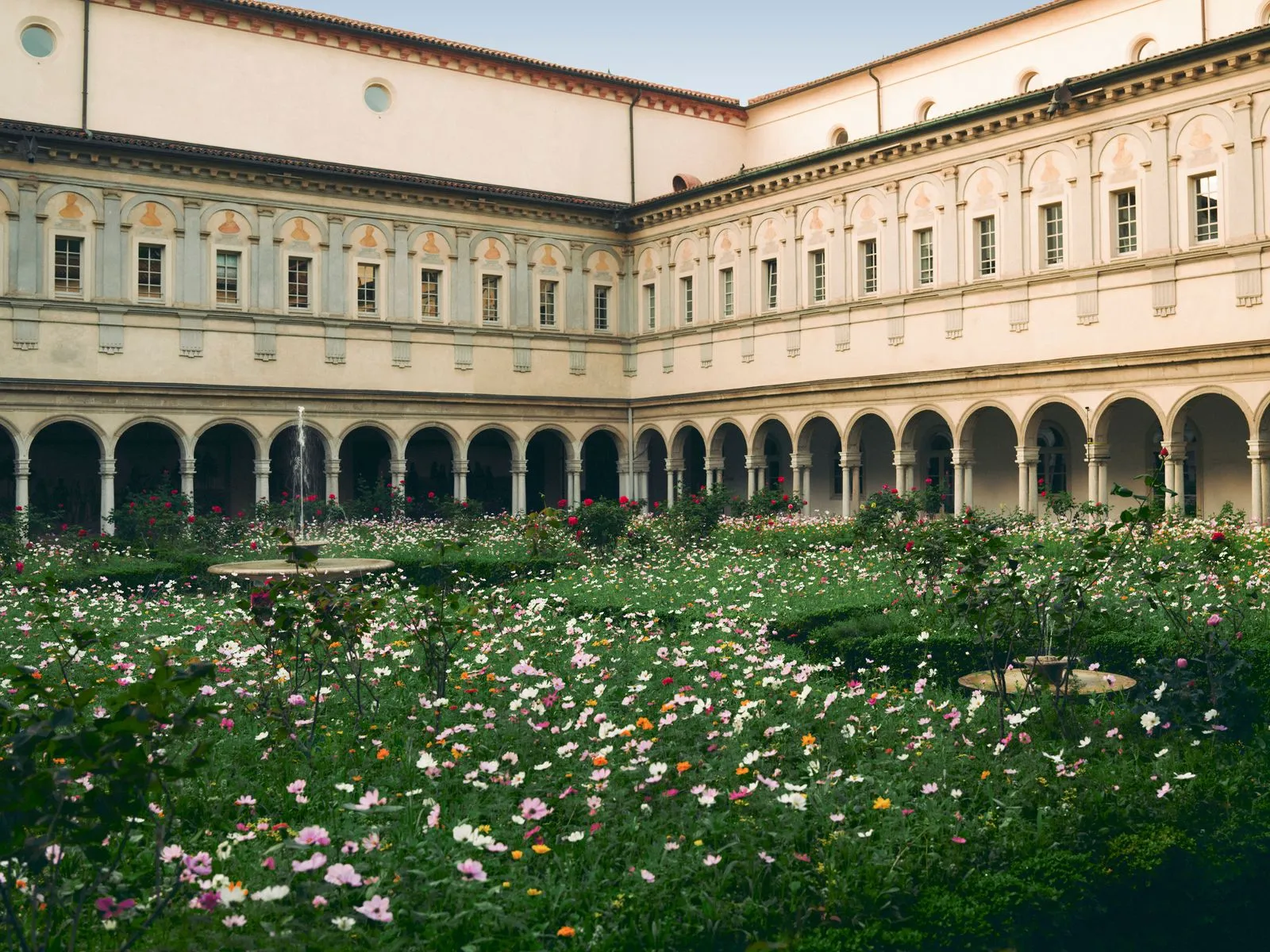 Historic courtyard with a fountain and a lush flower-filled garden surrounded by arcaded colonnades; cream-colored walls above.