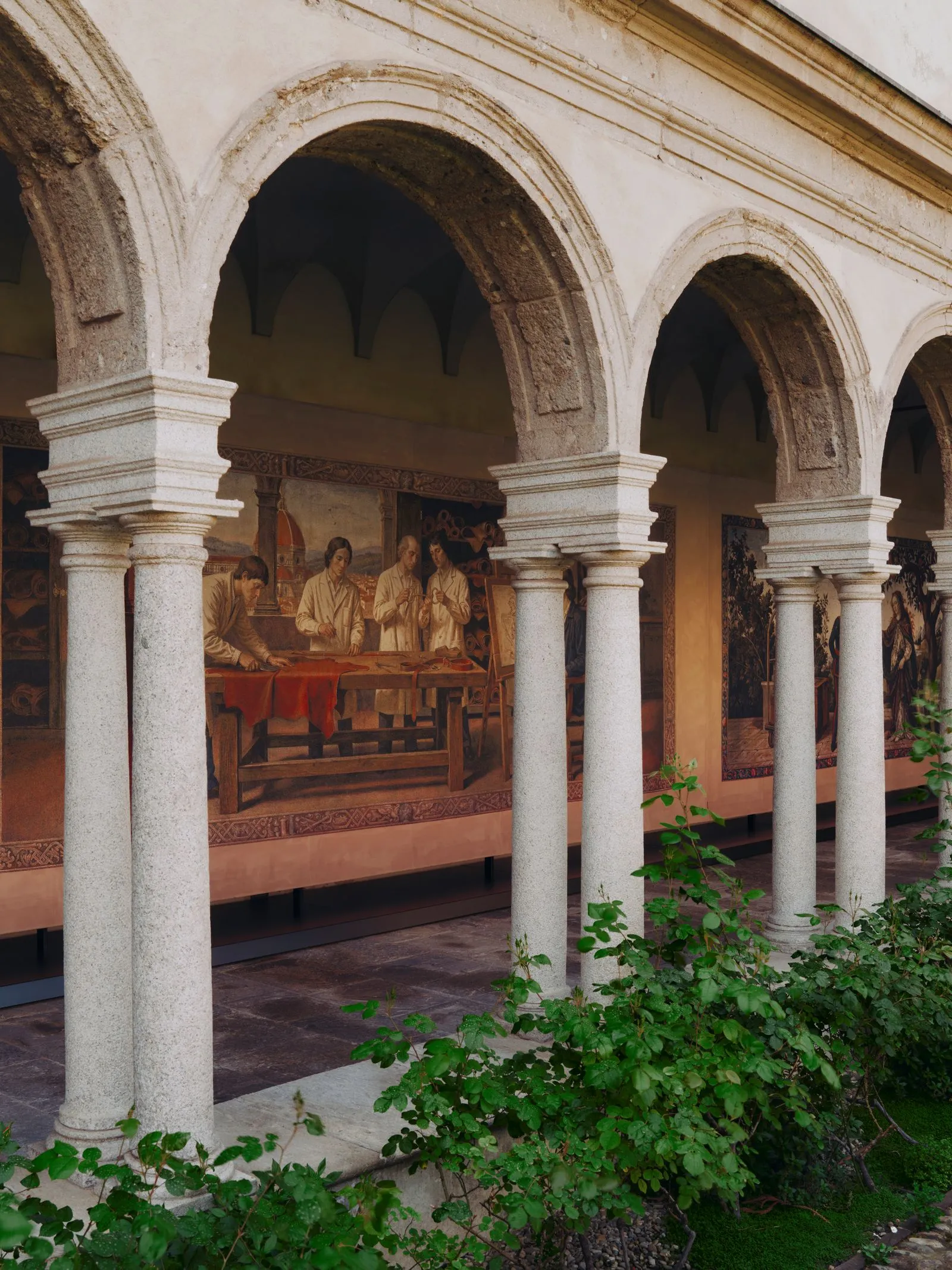 Stone arched colonnade with carved columns and frescoed wall paintings viewing into a courtyard; green plants in foreground.