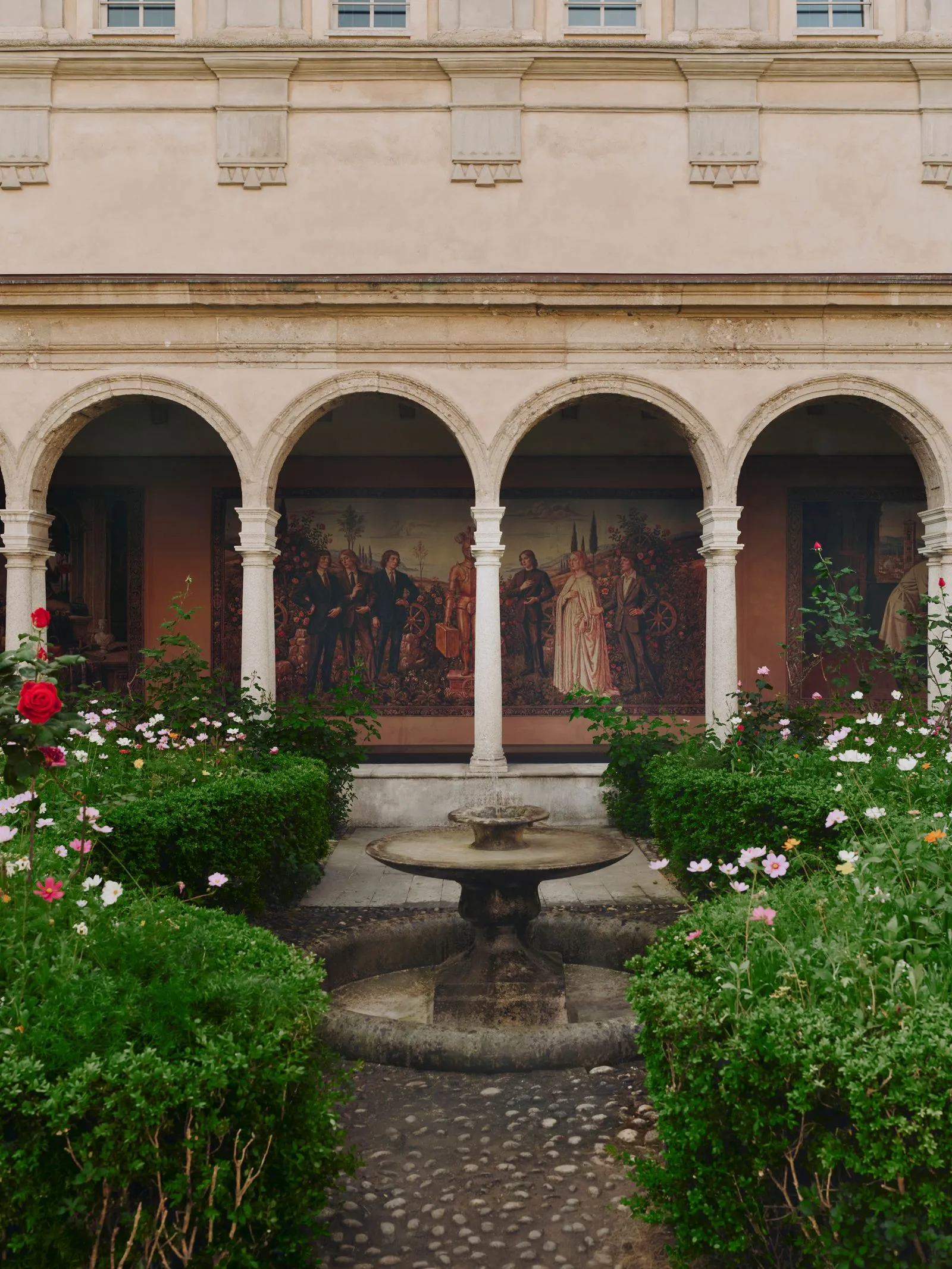 Courtyard with a row of stone arches and a central fountain, framed by blooming rose bushes and greenery.