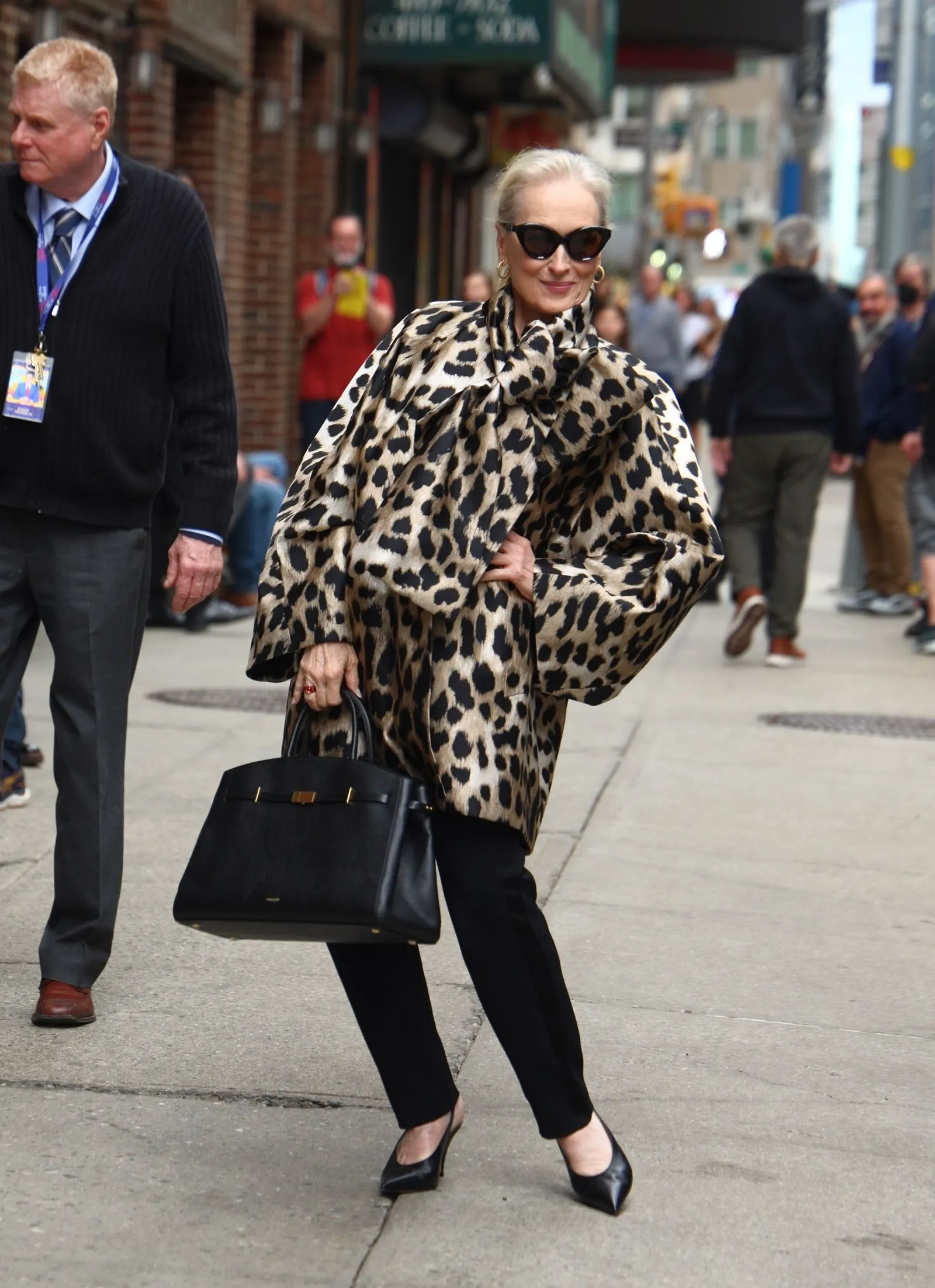 Elegant woman in a leopard-print cape, black pants and heels, wearing sunglasses and carrying a black handbag on a busy city street.
