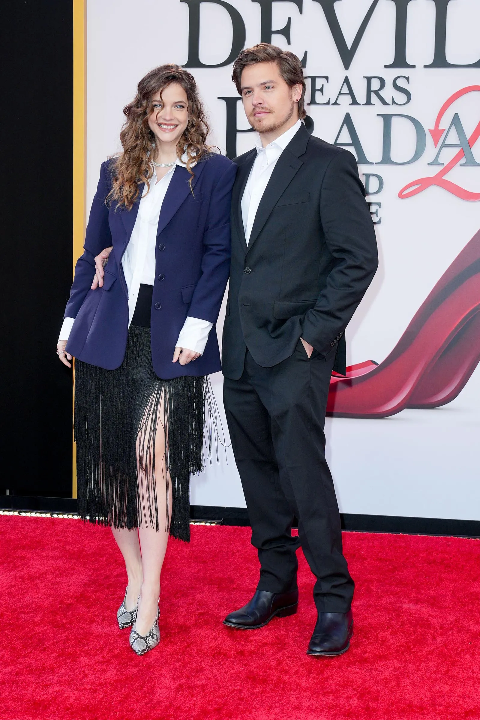 Two casually dressed adults pose on a red carpet, smiling at the camera with a decorative backdrop behind them.