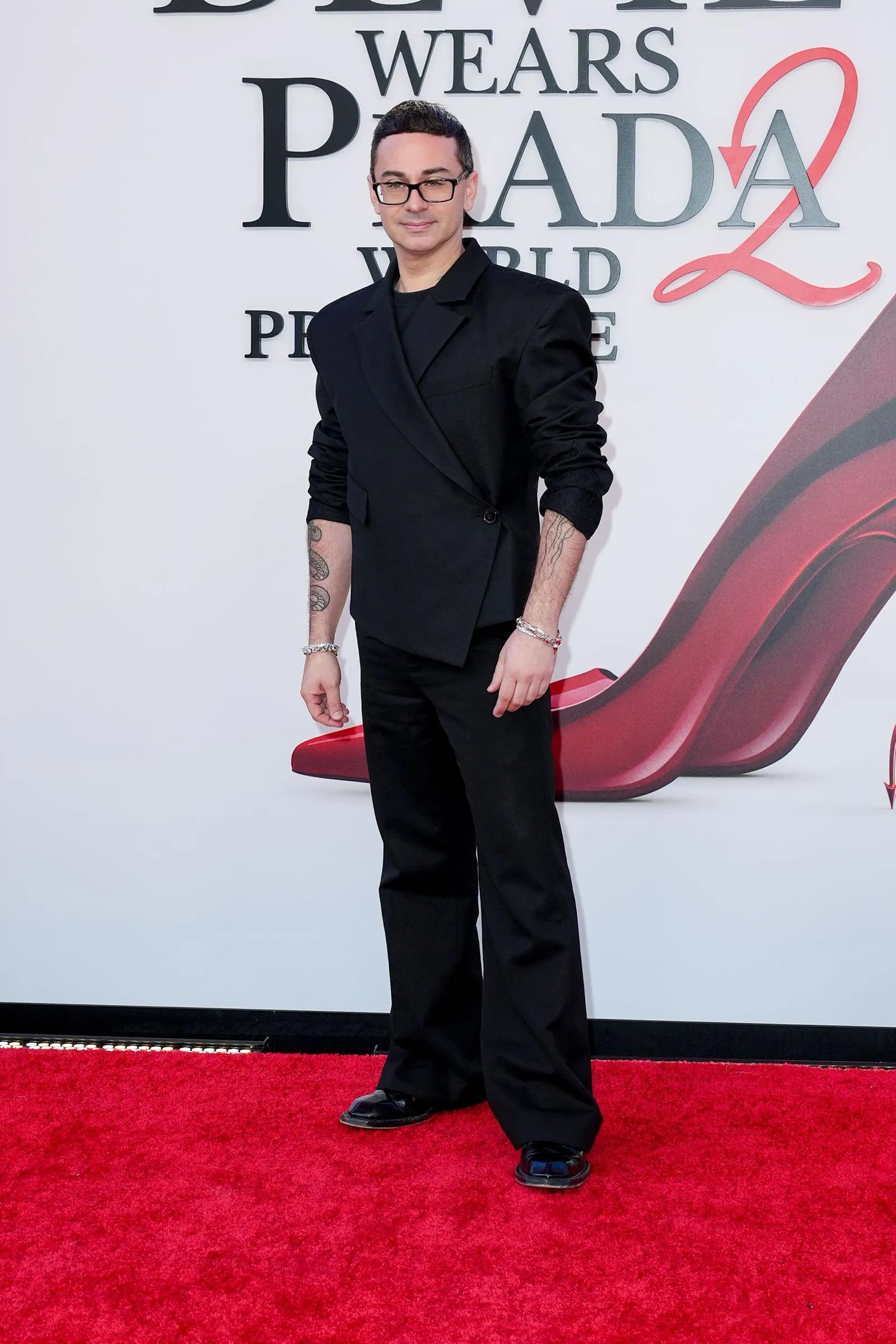 Man in a black suit with rolled sleeves and glasses posing on a red carpet at a fashion event; visible forearm tattoos and a backdrop featuring large Prada lettering.