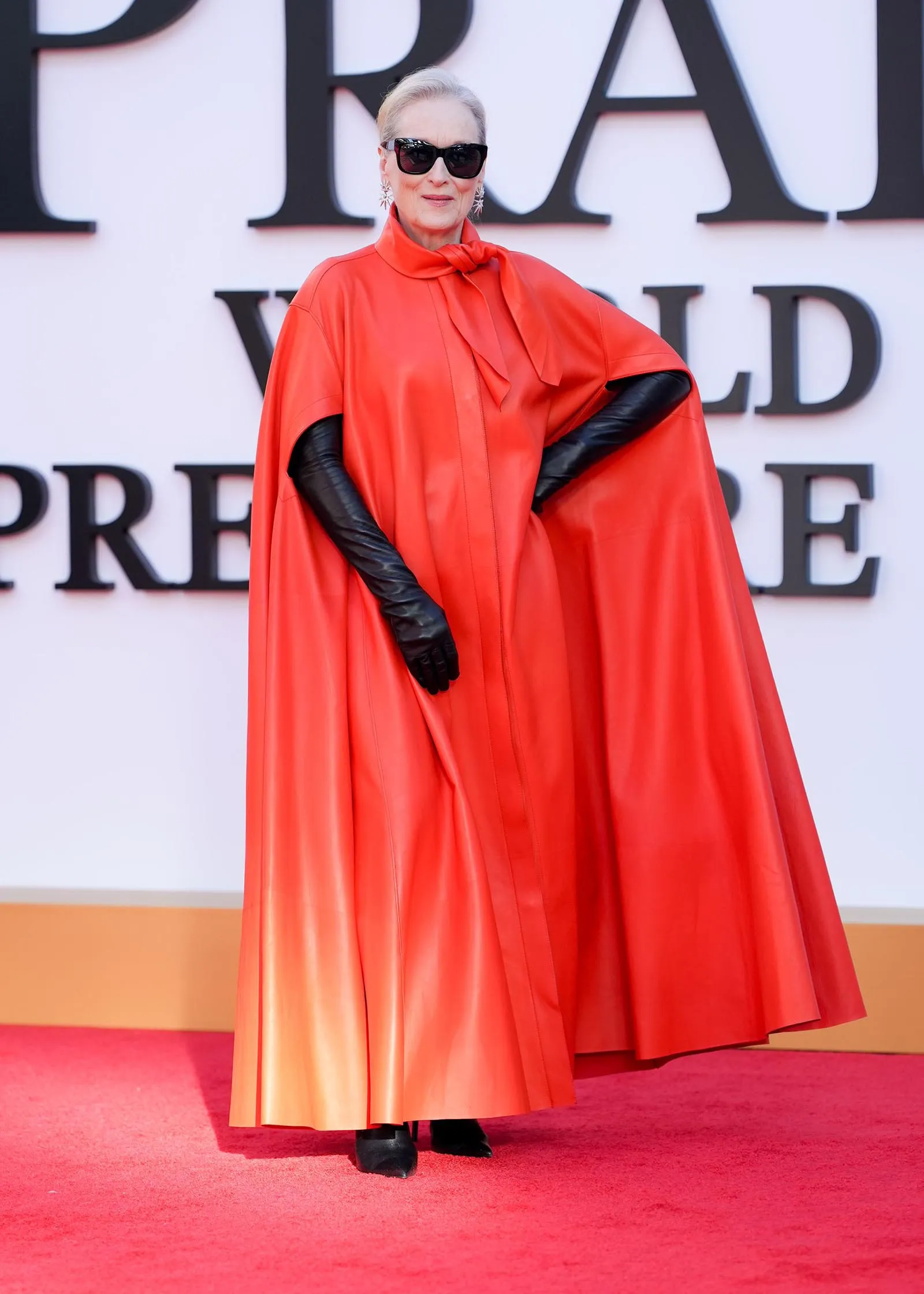 Person posing on a red carpet in a bright red cape dress, black opera gloves, dark sunglasses, and dangling earrings.
