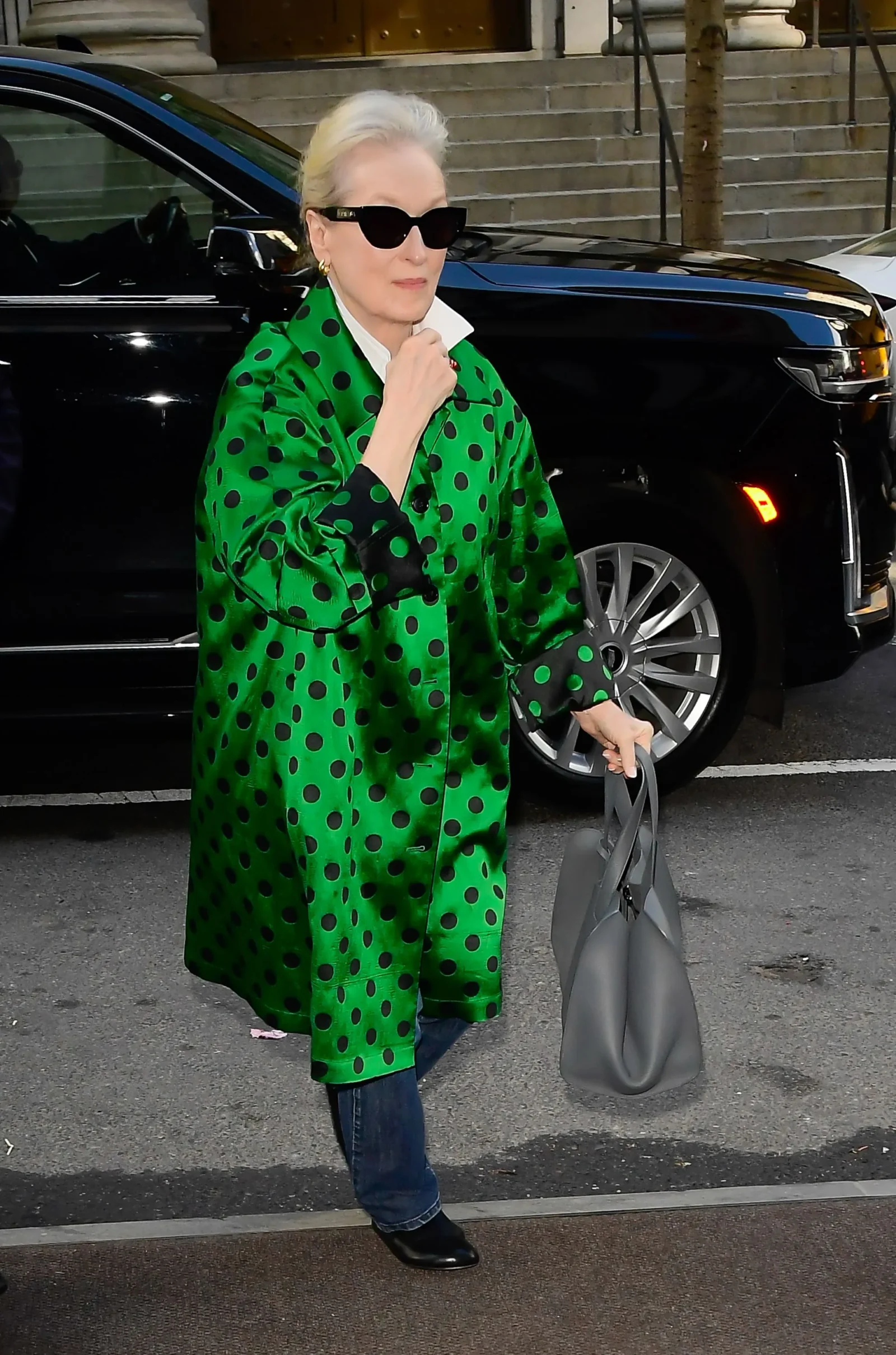 meryl streep in a bright green polka-dot coat with black dots, sunglasses, and blue jeans, standing beside a black car while holding a gray handbag.