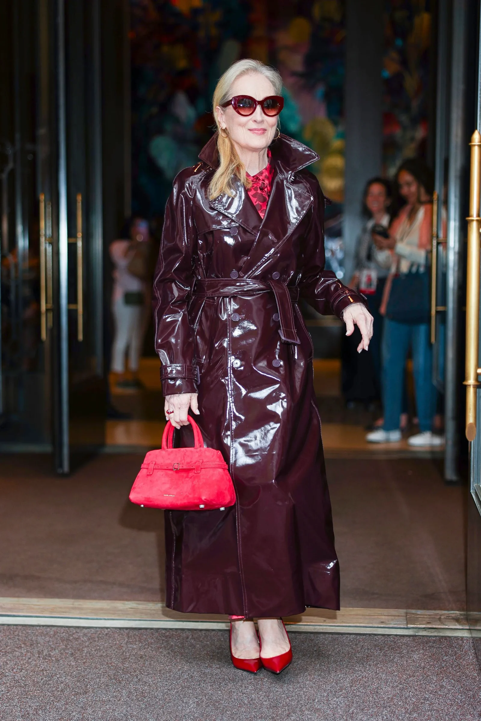 meryl streep in a shiny burgundy trench coat, red sunglasses, and red heels holds a bright red handbag at a doorway.