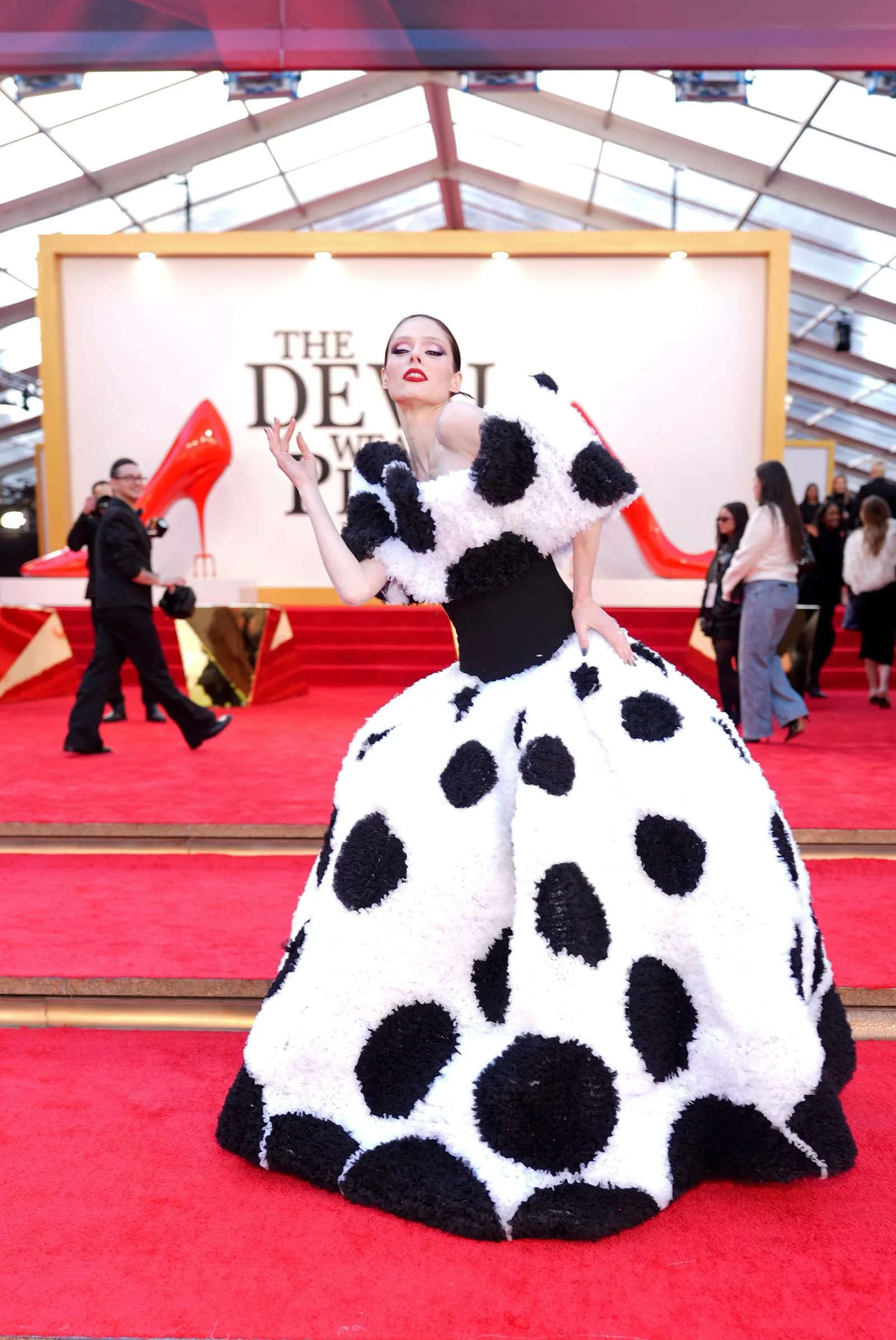 Model on a red carpet wearing a dramatic white ballgown with large black polka dots, posing with a raised hand.