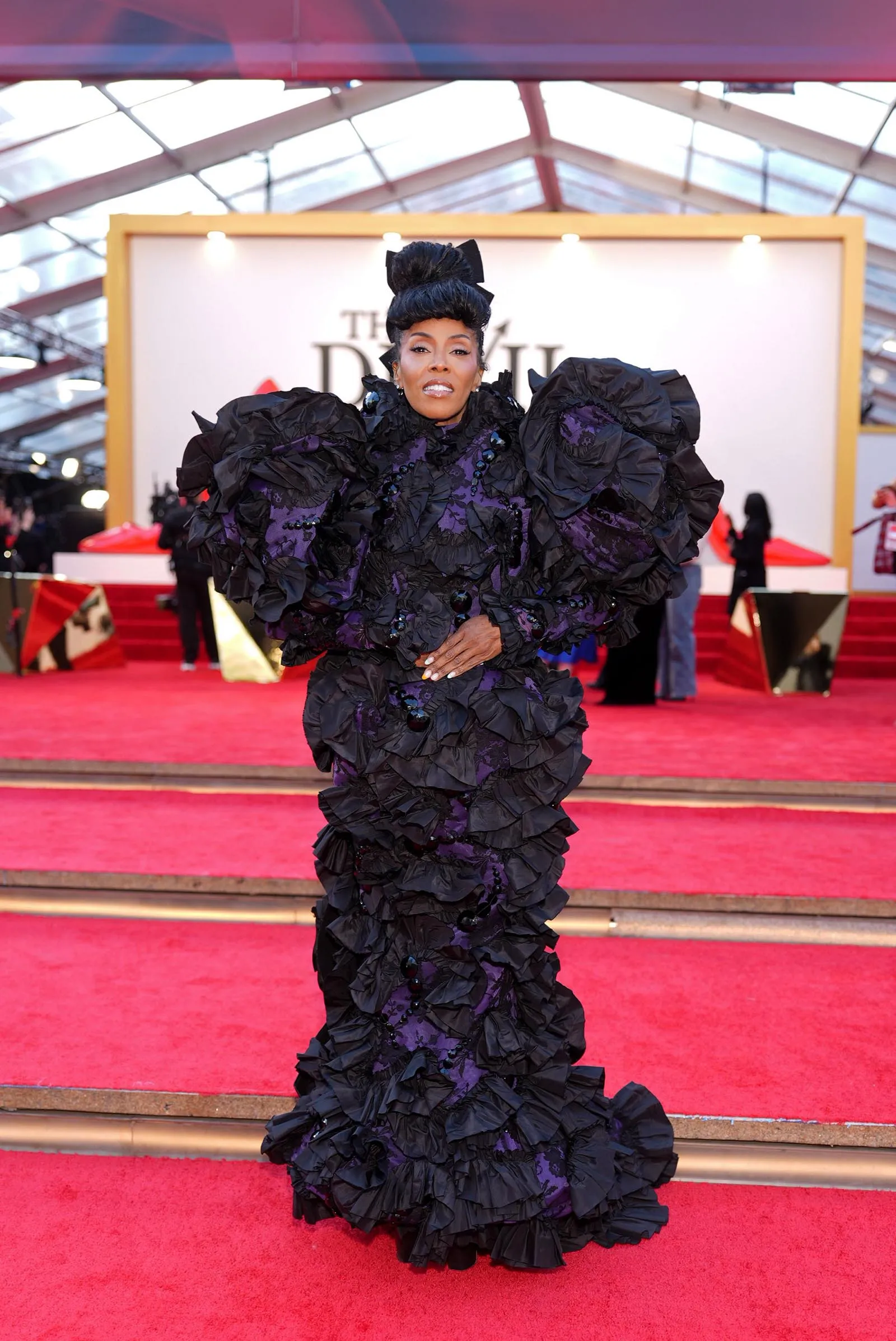 Woman on a red carpet wearing a dramatic black and purple ruffled gown with oversized sleeves, posing for photos.