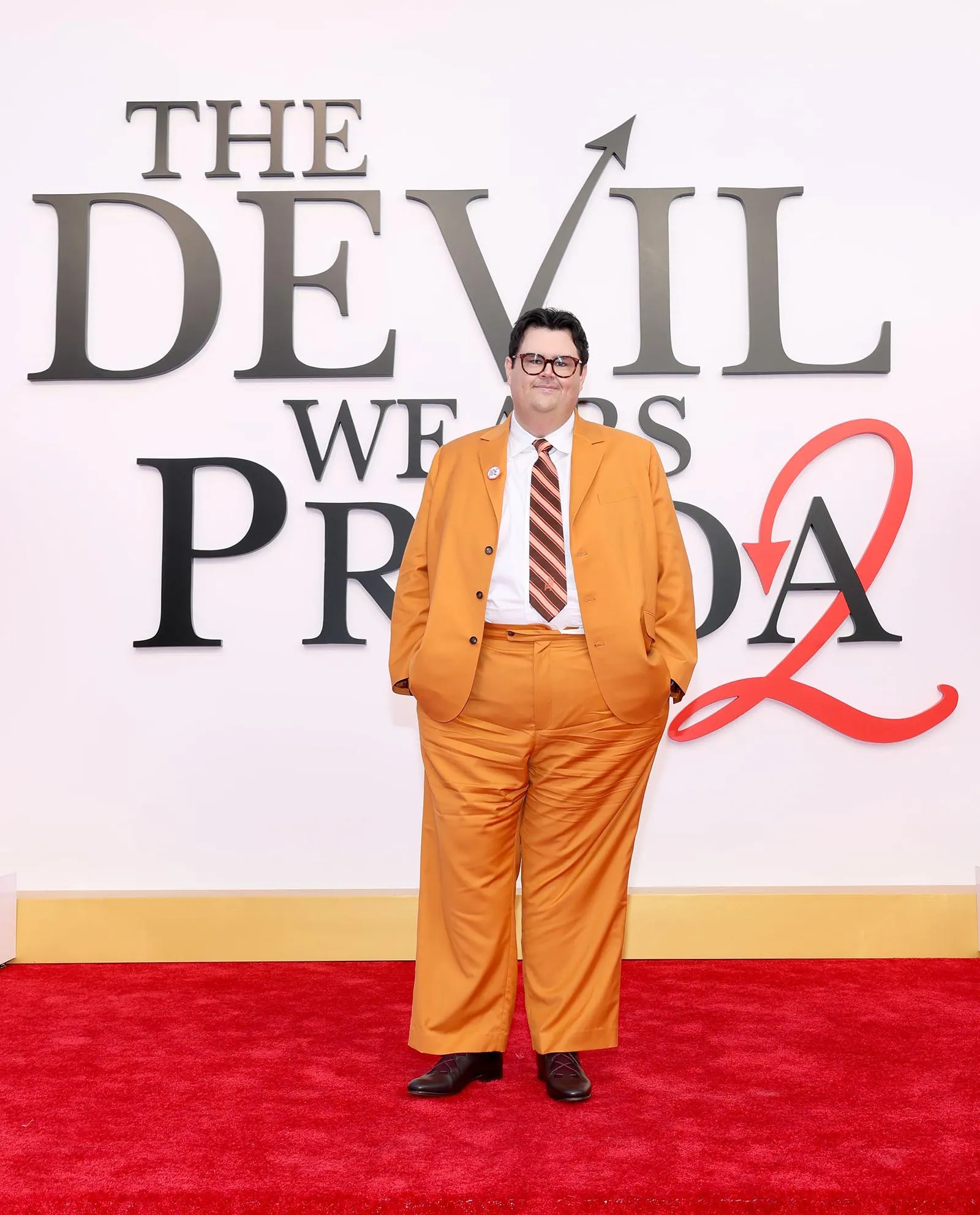 Man in an orange suit posing on a red carpet in front of a large backdrop that reads 'THE DEVIL WEARS PRADA'.