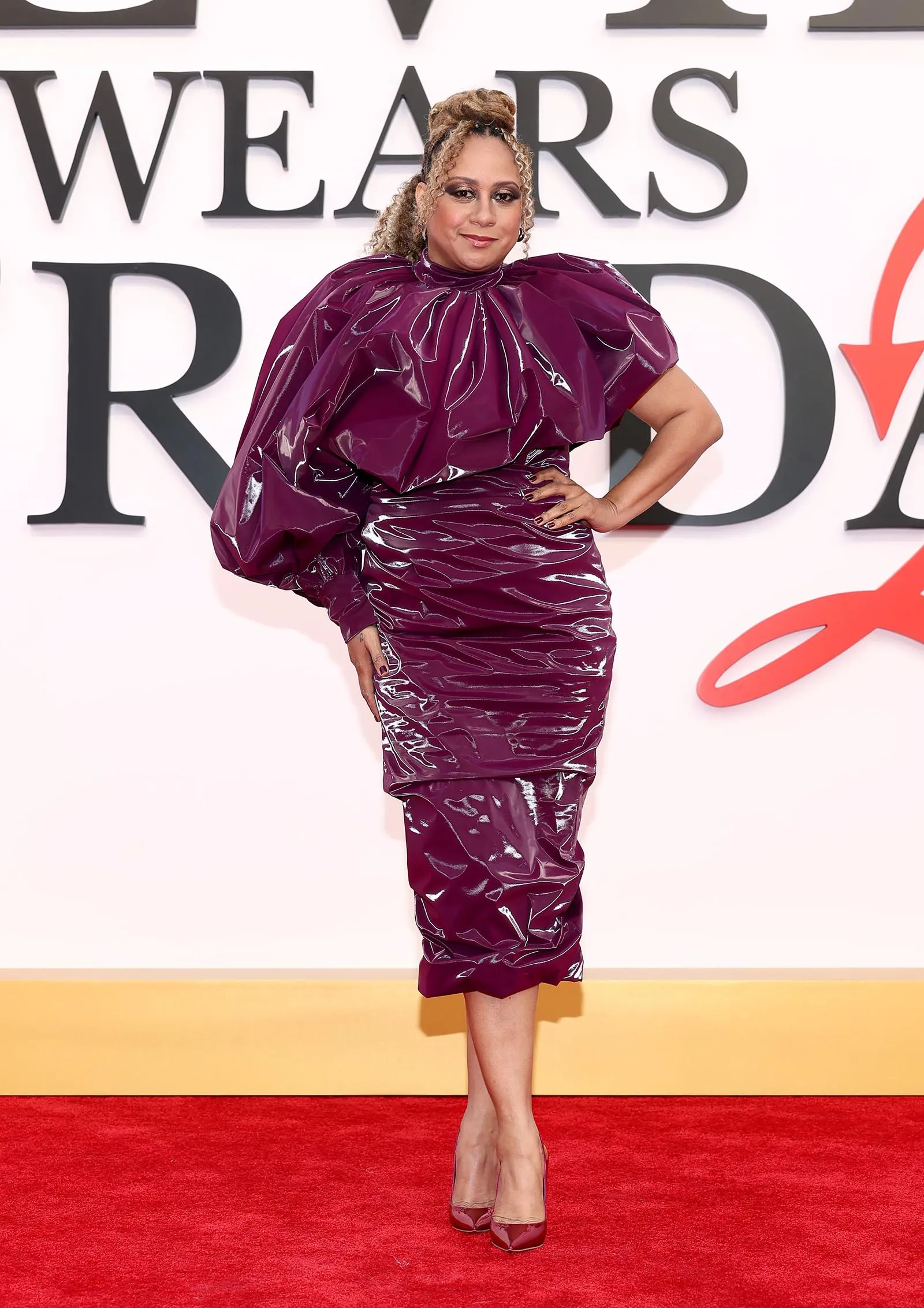 Woman on a red carpet wearing a shiny plum-purple dress with dramatic shoulder details and matching heels.