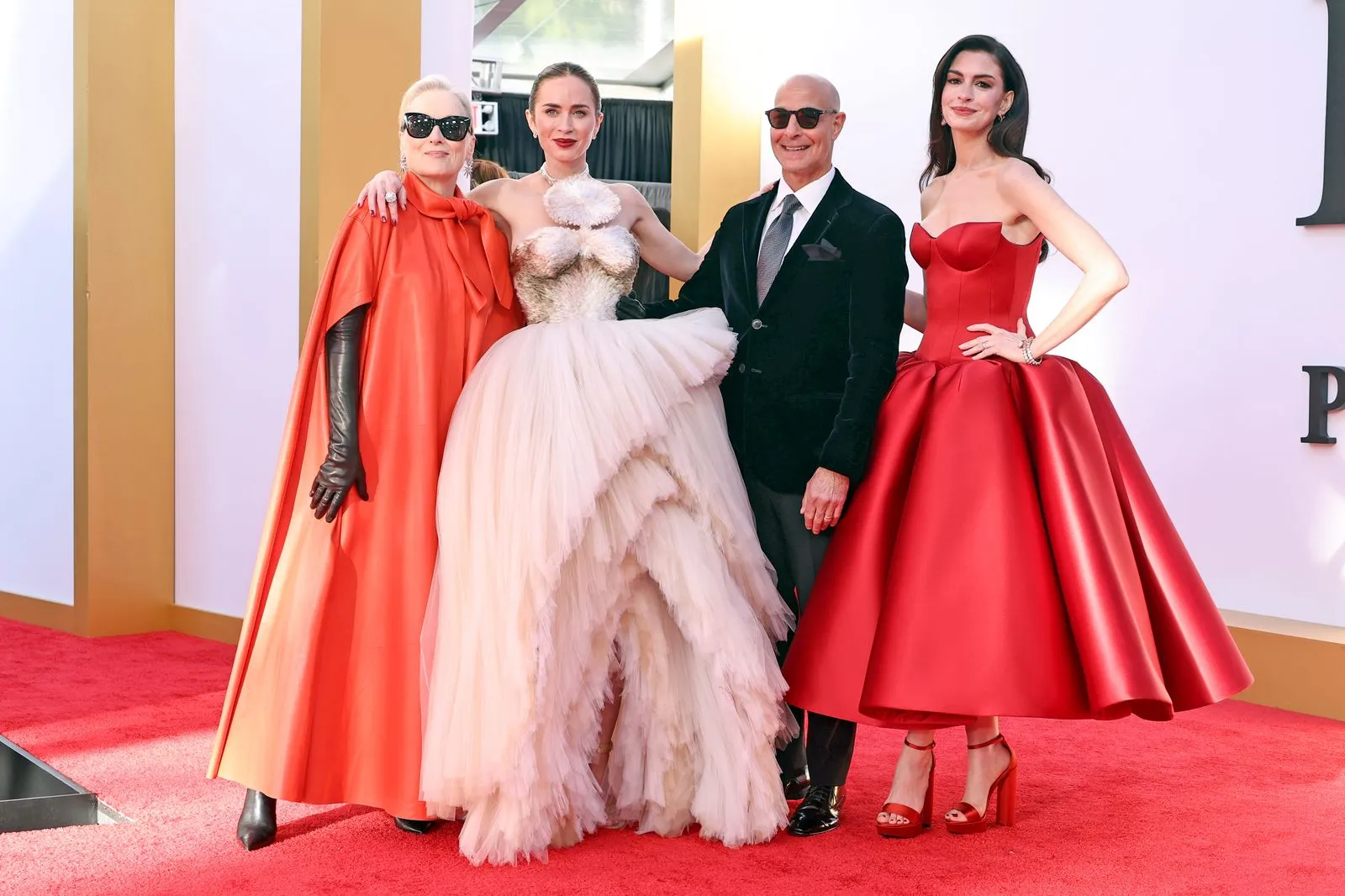 Four adults pose on a red carpet: woman in orange cape dress and gloves, woman in pale pink fluffy gown, man in dark suit with sunglasses, woman in red strapless ballgown.