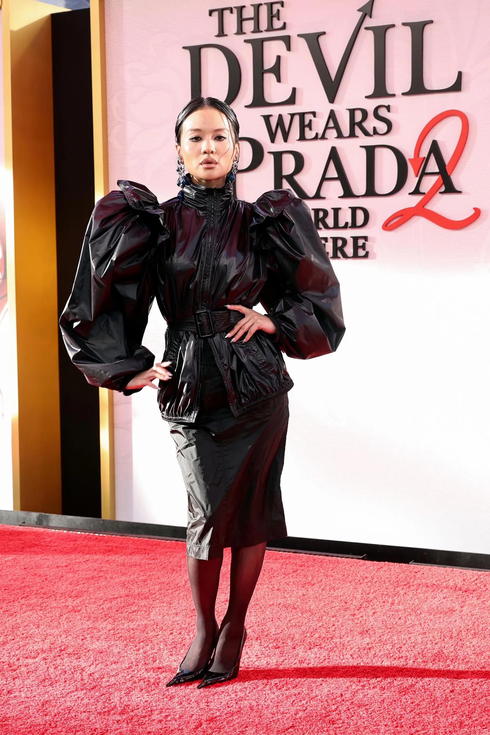 Woman posing on a red carpet in a shiny black puff-sleeve outfit, The Devil Wears Prada backdrop behind her.