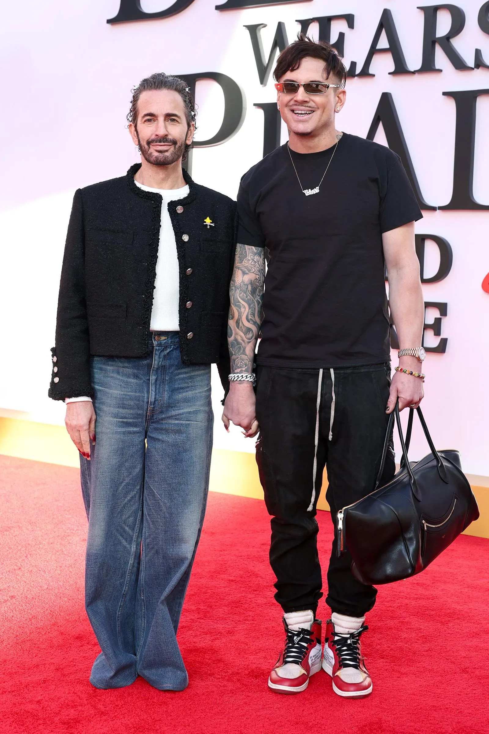 Two men posing on a red carpet at an event; one in a black textured jacket and jeans, the other in a black t‑shirt with tattoos and a black bag.