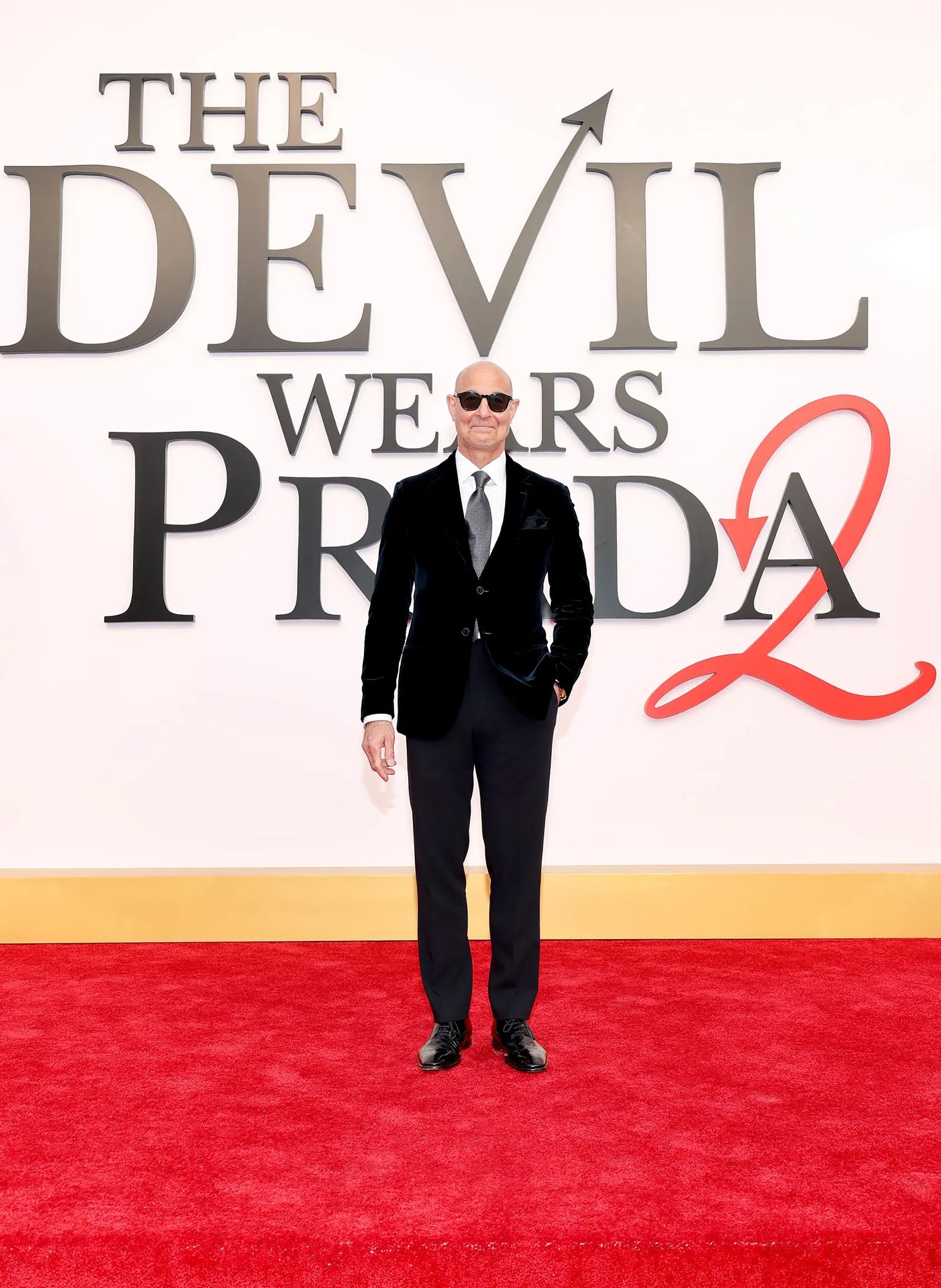 Man in a dark suit and sunglasses posing on a red carpet with a The Devil Wears Prada backdrop behind him.