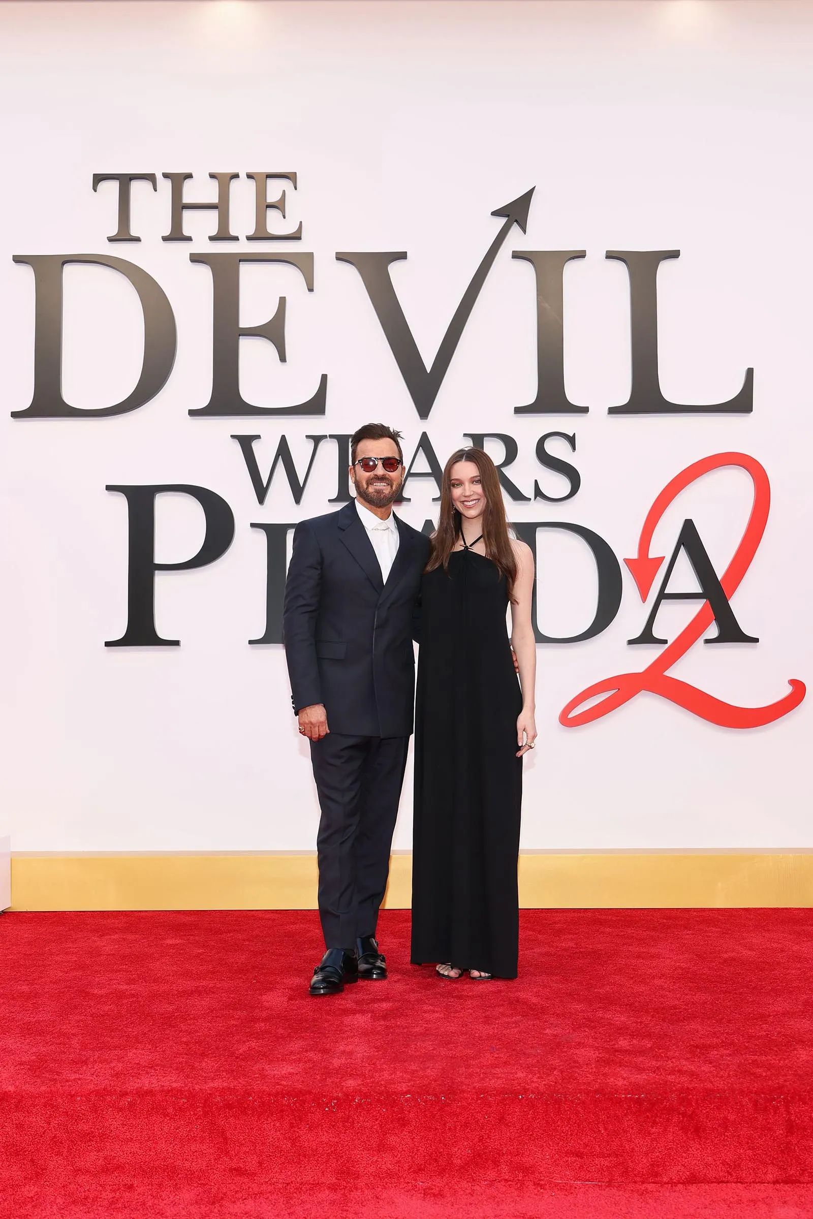 Couple posing on a red carpet in front of a large 'The Devil Wears Prada' backdrop.