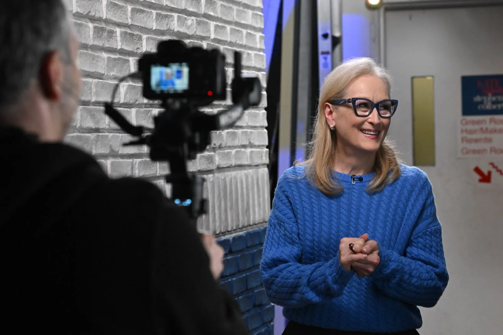 Woman in a blue knit sweater smiles while being filmed in a studio interview setup, with a brick wall backdrop and a camera in the foreground.