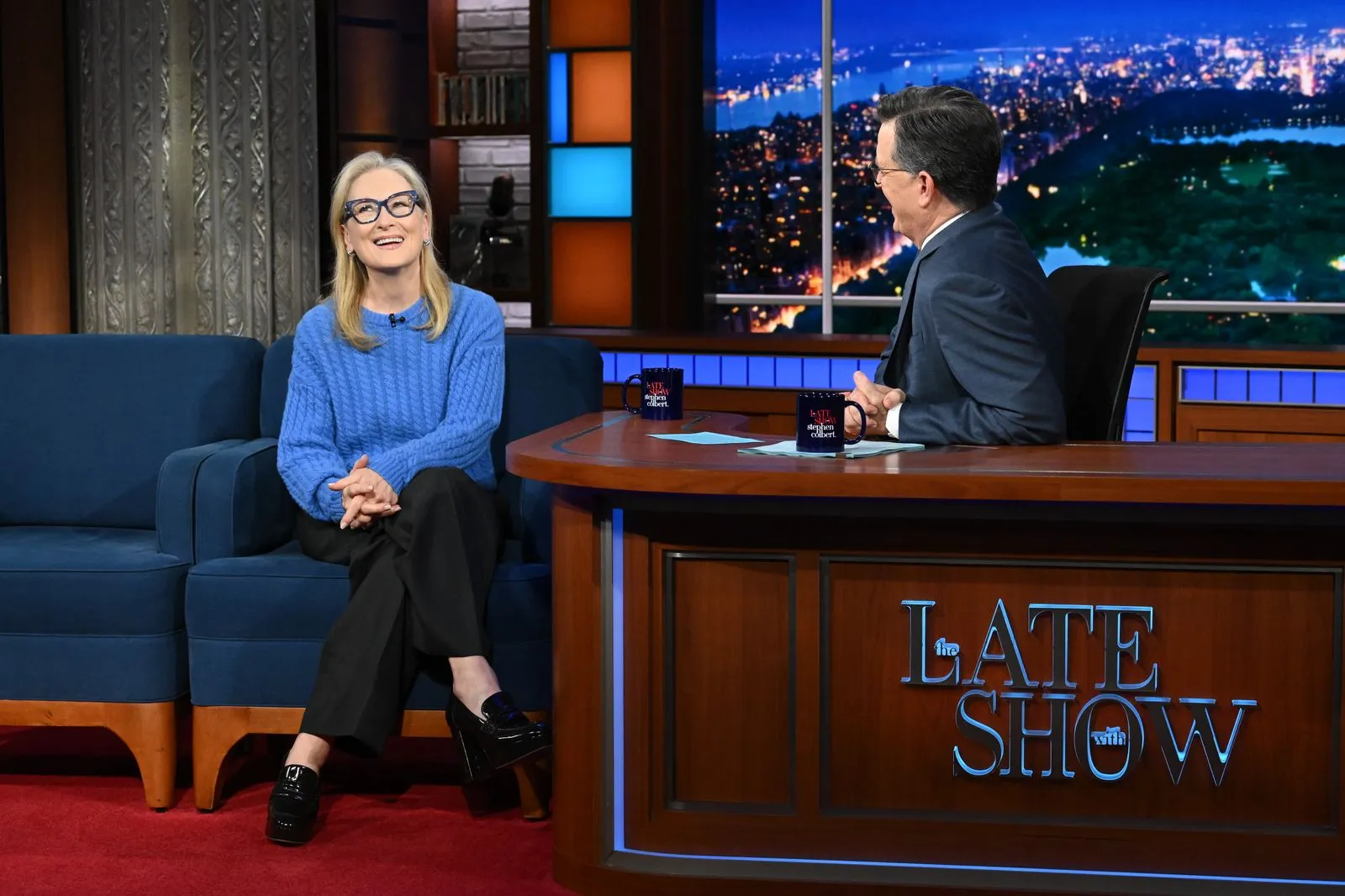 Talk-show interview scene: a woman in a blue knit sweater sits on a blue sofa, smiling, while the host sits at a wooden desk with papers and mugs nearby, city lights backdrop behind them.