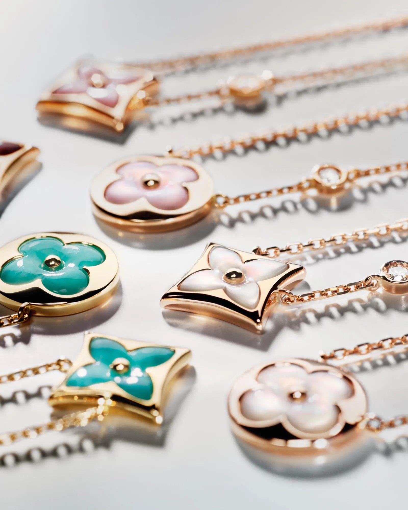 Assorted gold pendant necklaces with enamel flower designs in pink, teal, and white on a reflective surface.