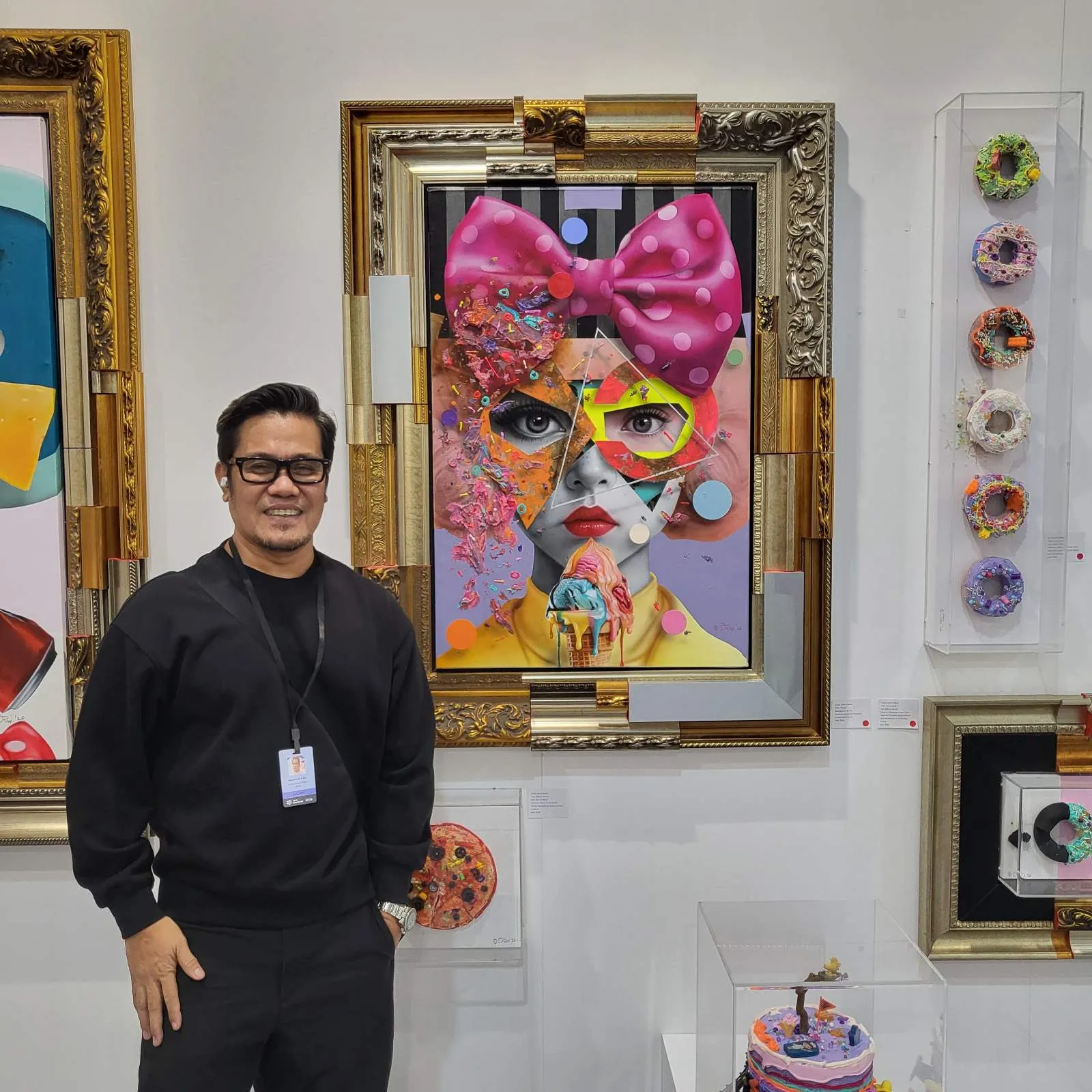 Man in black with a lanyard posing in front of a colorful framed portrait of a girl with a pink polka-dot bow in the center of the image.