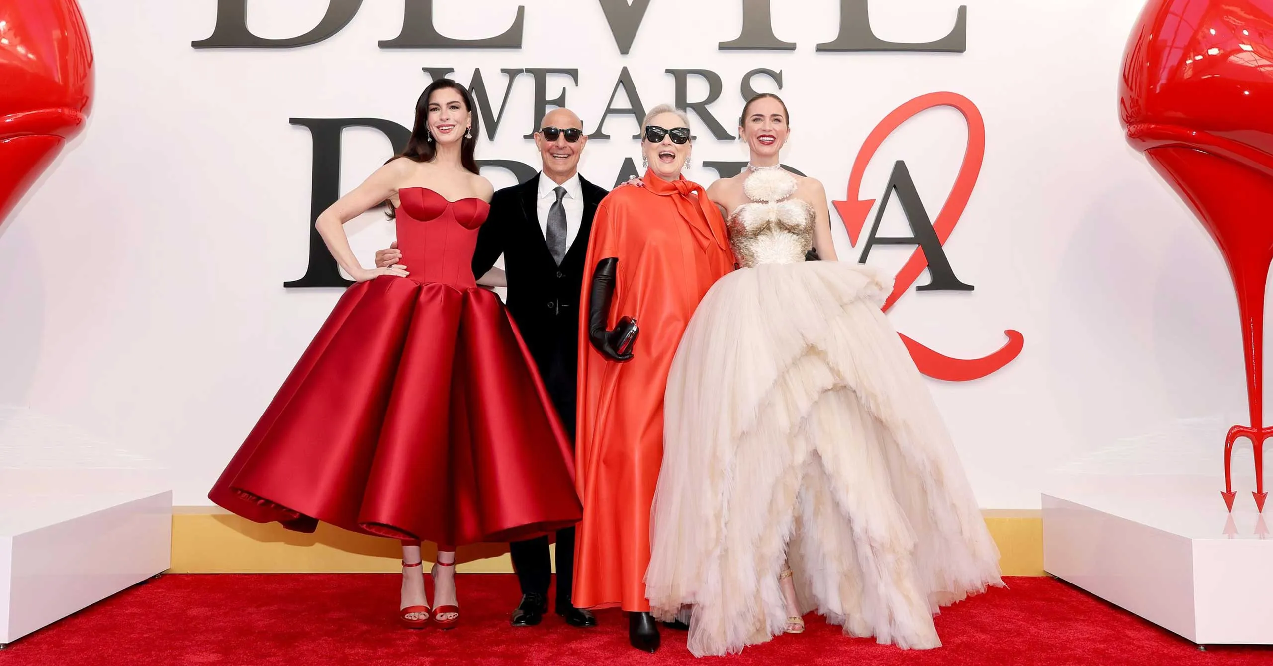 Four people pose on a red carpet in front of a white backdrop with large black letters, wearing elegant evening gowns and a suit.