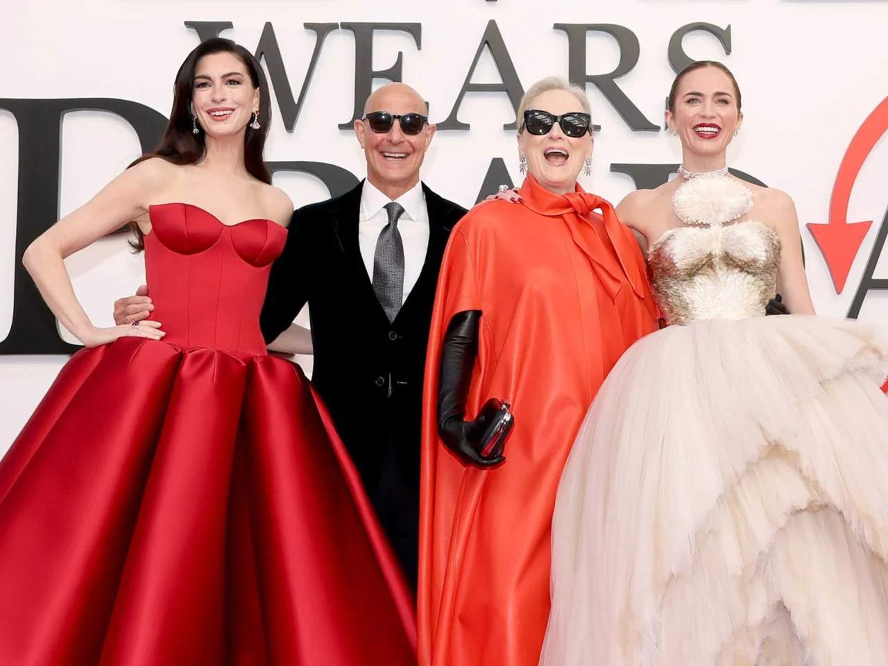 Four people pose on a red carpet in front of a white backdrop with large black letters, wearing elegant evening gowns and a suit.