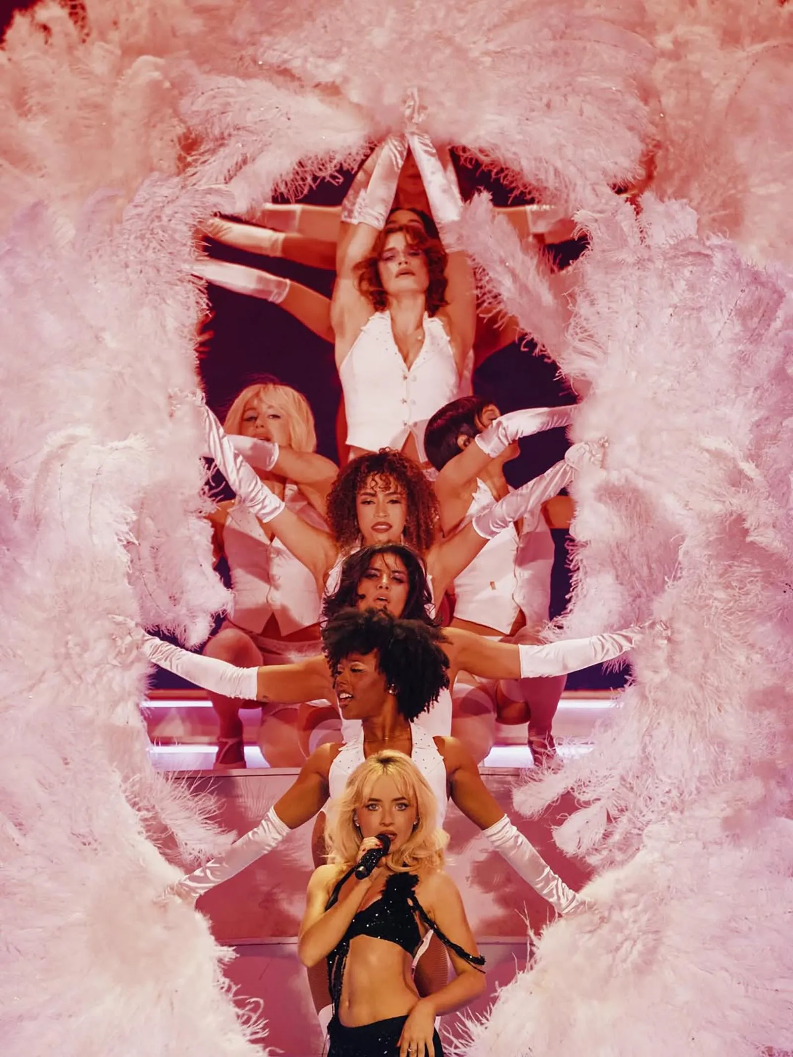 Blonde female singer with a microphone performs center stage, surrounded by dancers in white behind a pink feathered framing arch.