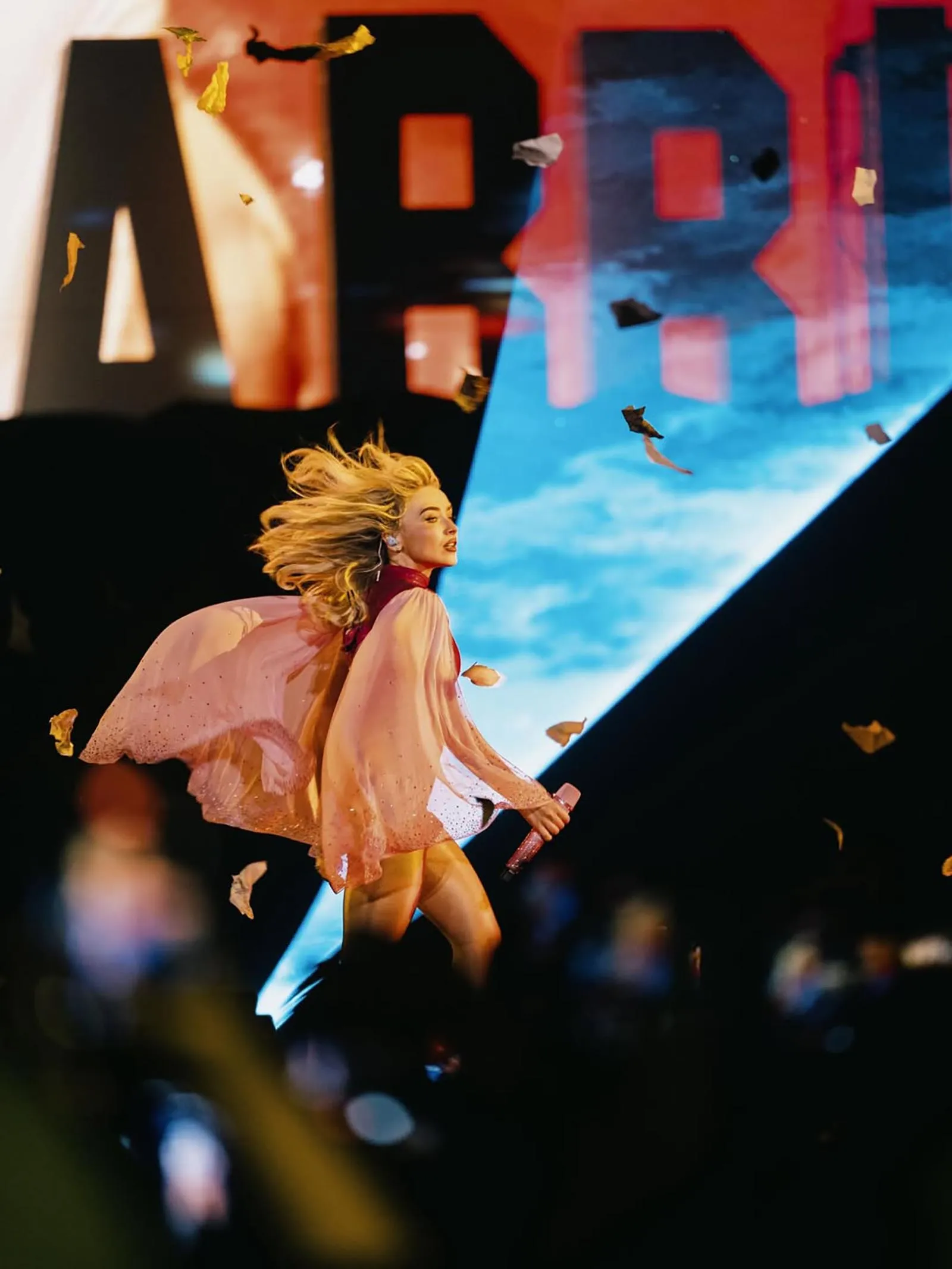 Female pop singer in a pink flowing cape mid-performance on stage, microphone in hand, confetti falling around.