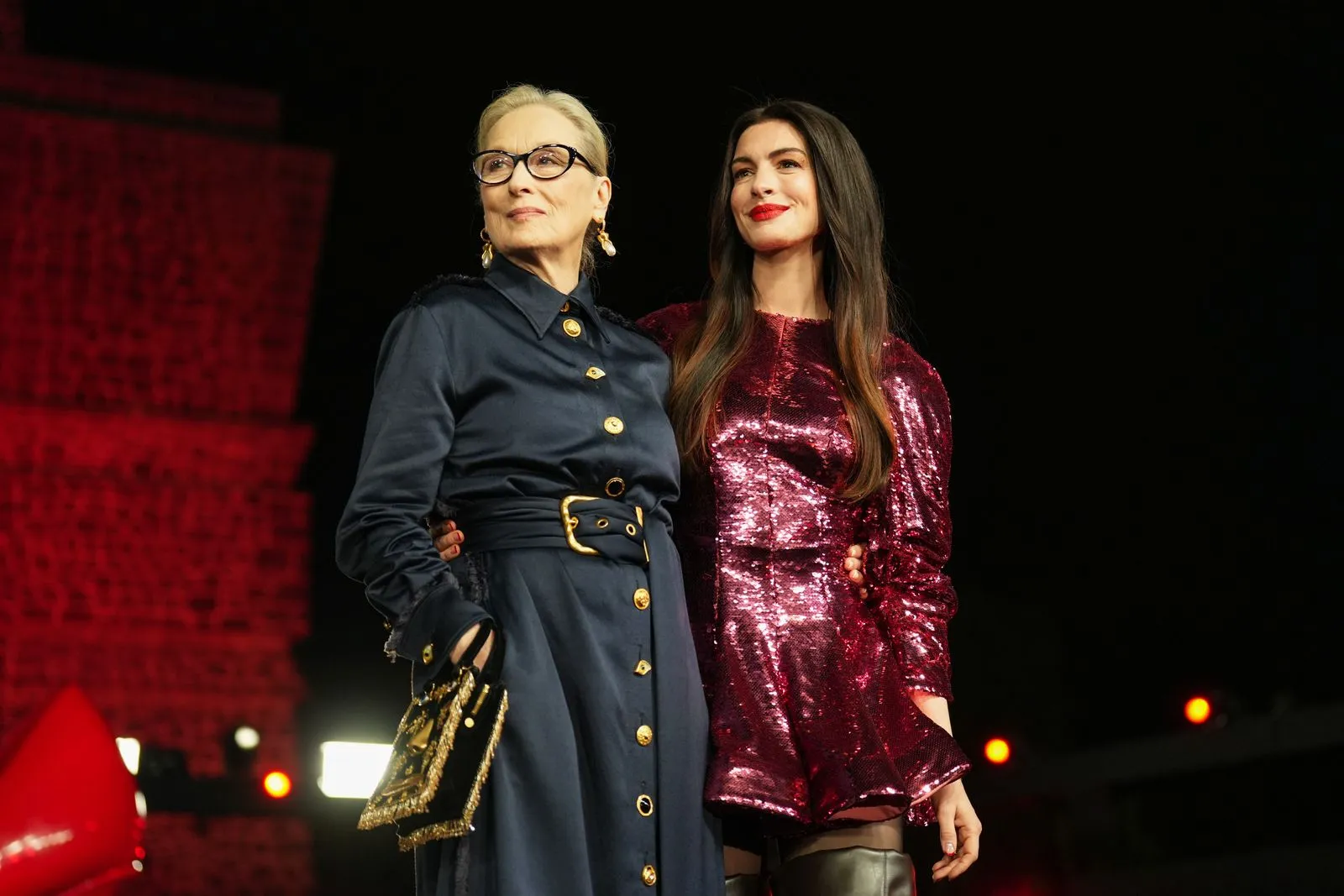 Two women posing on a dark stage: one in a navy belted coat with gold buttons and glasses, the other in a pink sequin dress with long dark hair.