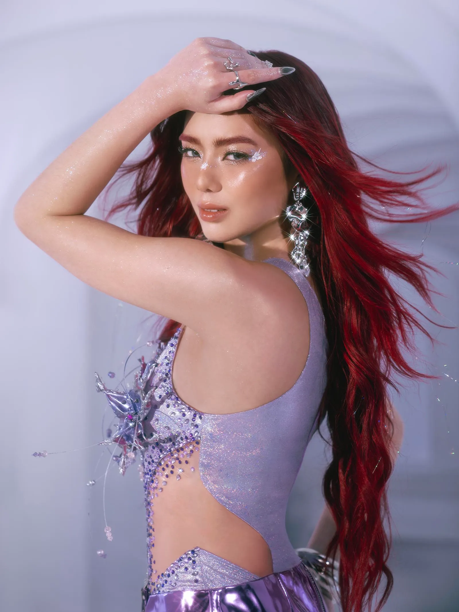 Portrait of a woman with long red hair wearing a lavender sparkly outfit, glittery eye makeup, and large crystal earrings, posing with her hand over her forehead.