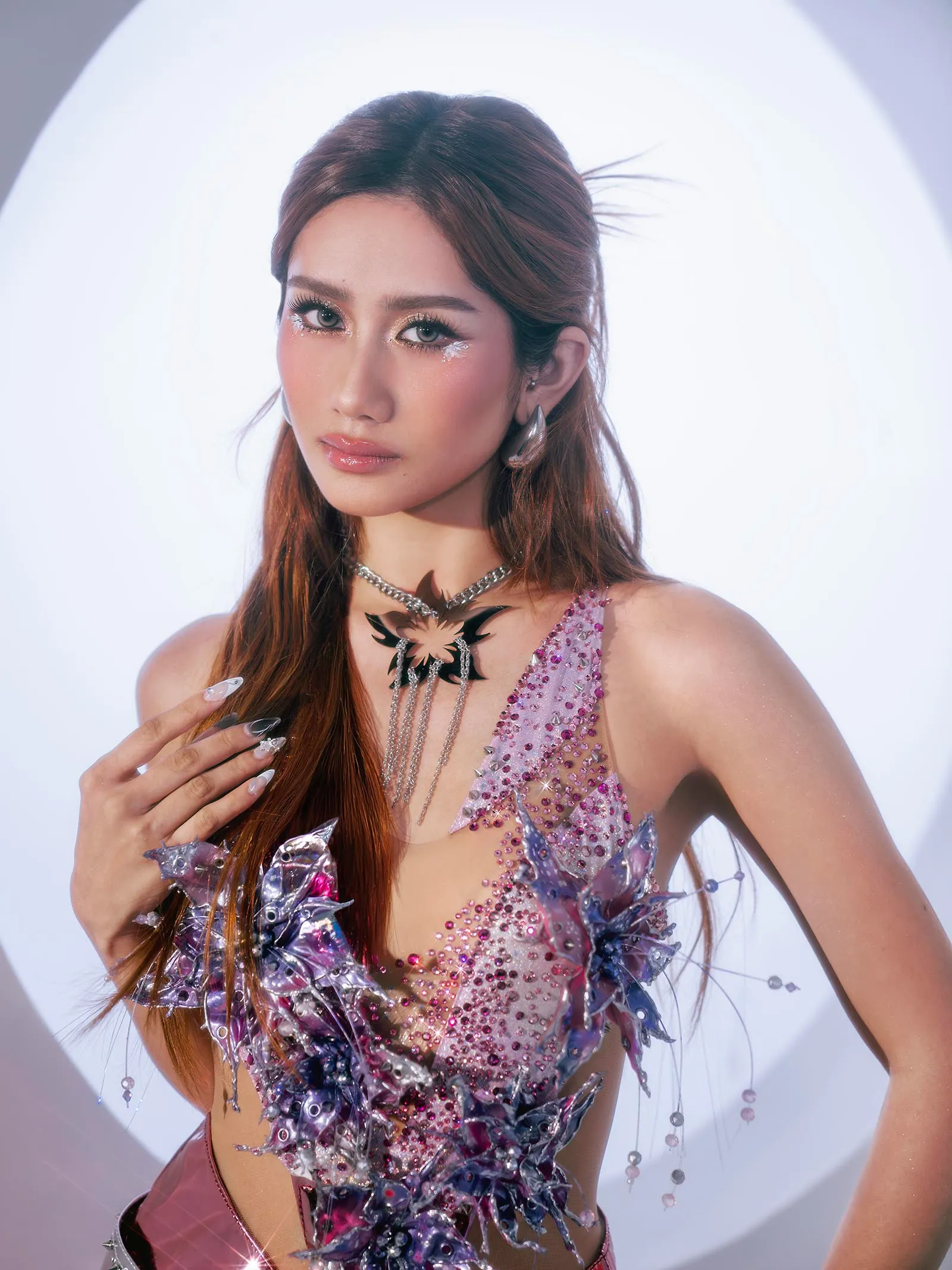 Portrait of a young woman in a pink rhinestone costume with glitter makeup, against a bright white backdrop.