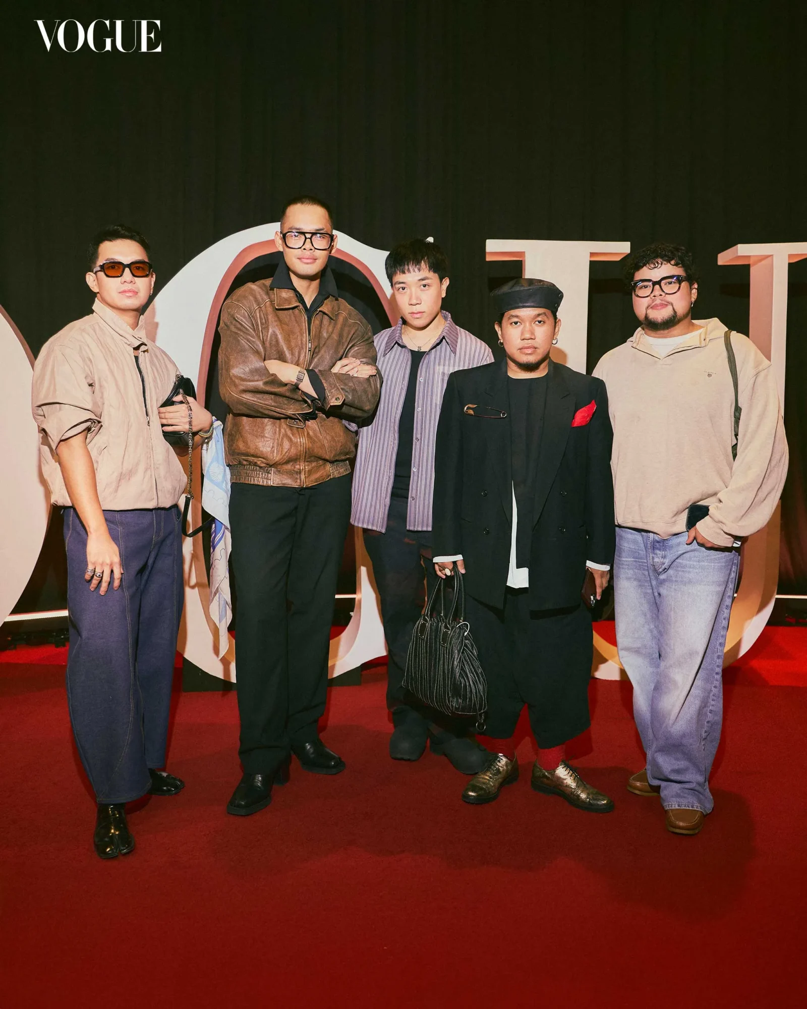 Five men pose on a red carpet at a Vogue event, wearing retro-inspired outfits with sunglasses, against a dark backdrop with large letters behind them.