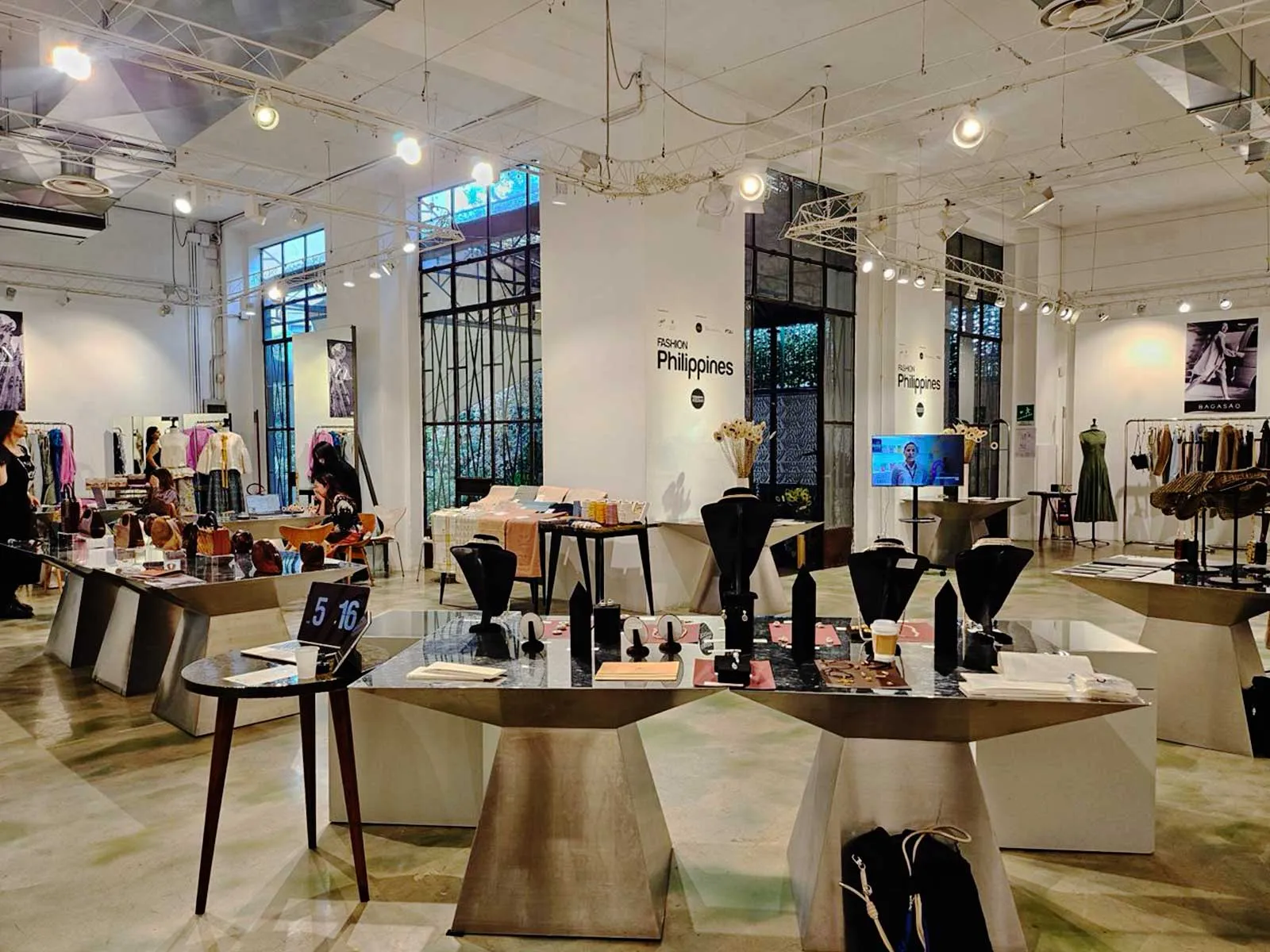 Bright fashion expo hall with clothing racks, jewelry displays on marble tables, and mannequins under industrial lighting; 'Fashion Philippines' signage on white walls.