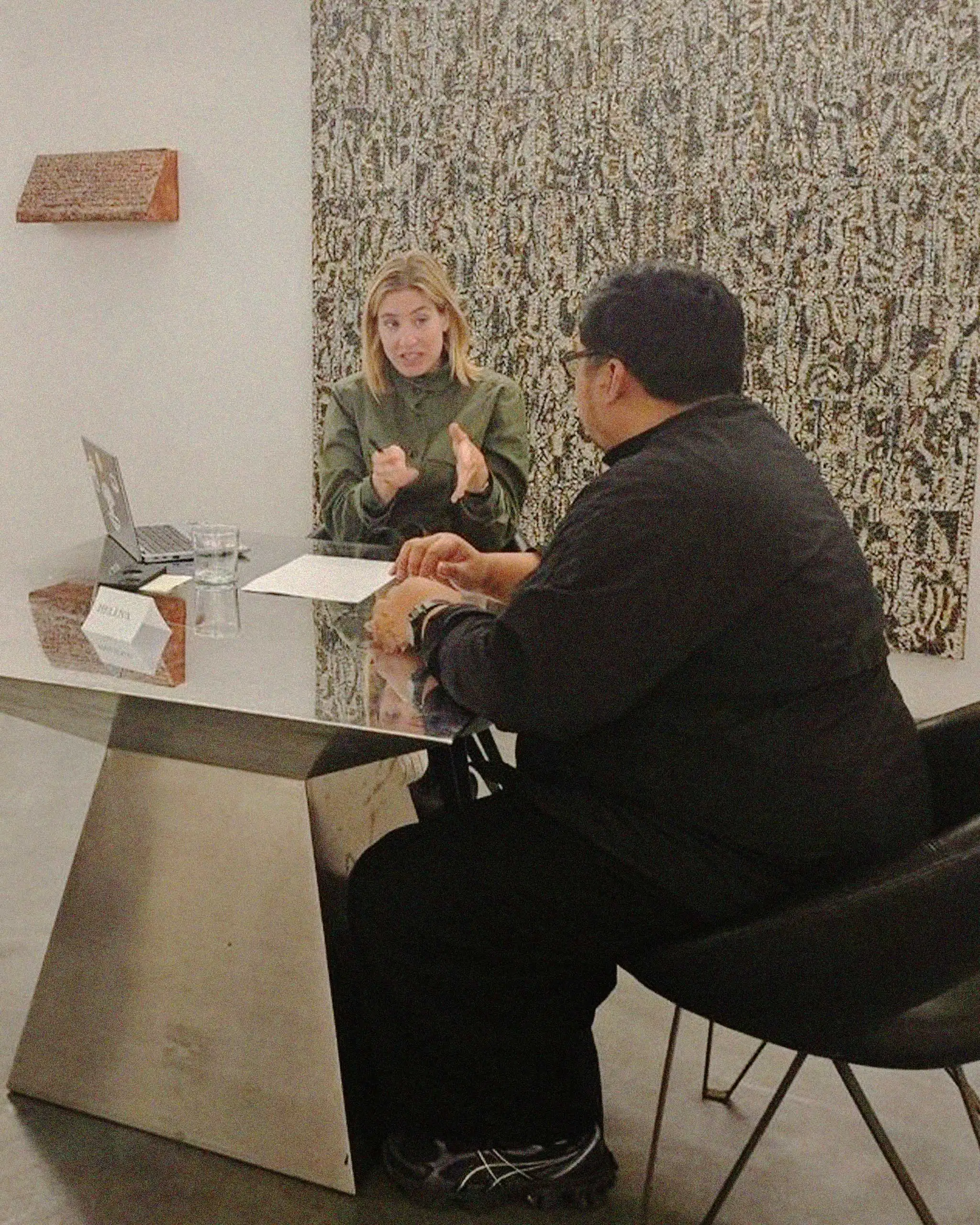 Two people sit across a glass desk; a blonde woman gestures while explaining to a man in a black jacket, with a laptop and papers on the desk.