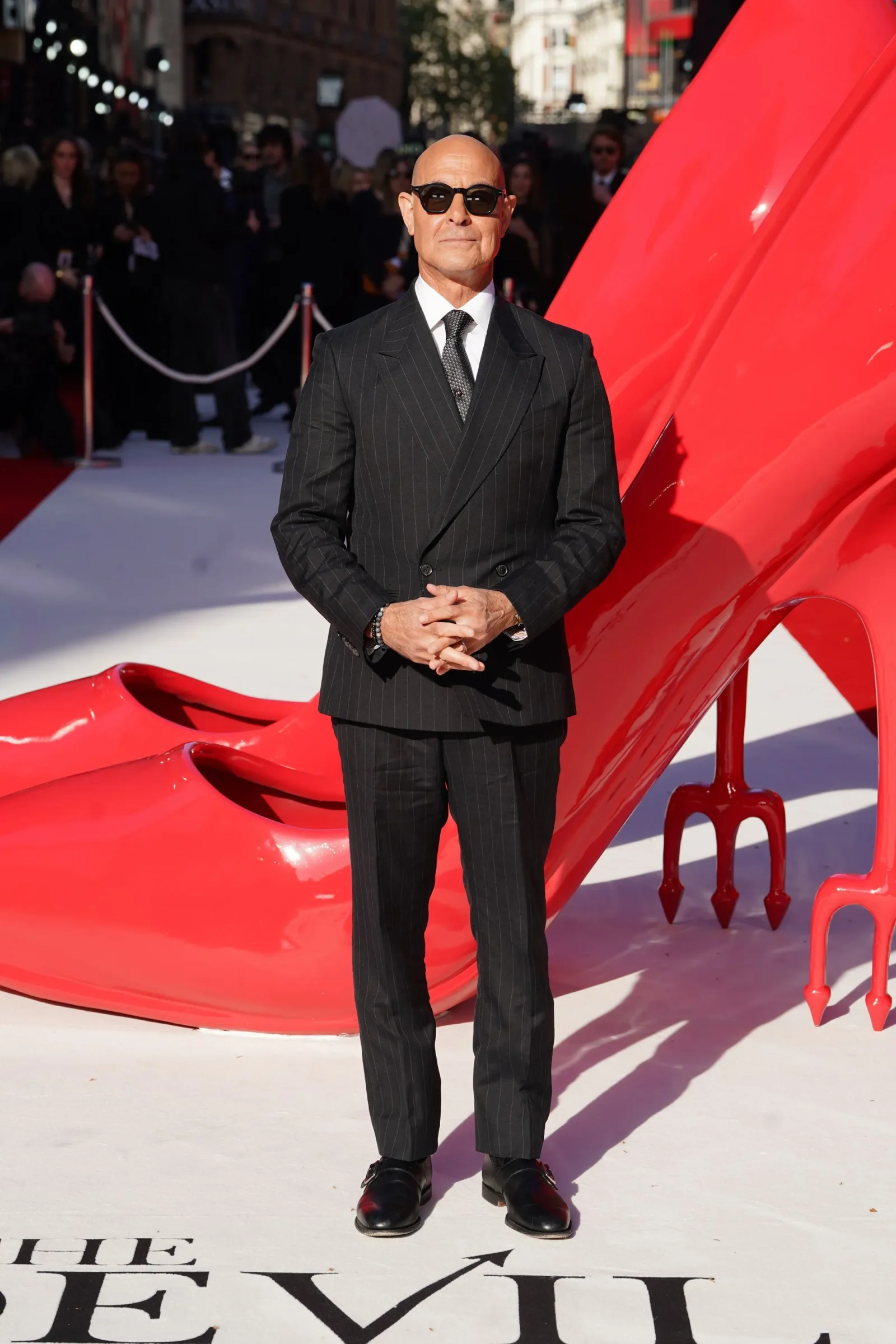 Man in a dark pinstripe suit and sunglasses stands with hands clasped on a white/red carpet beside a glossy red sculpture at a crowd-filled event.