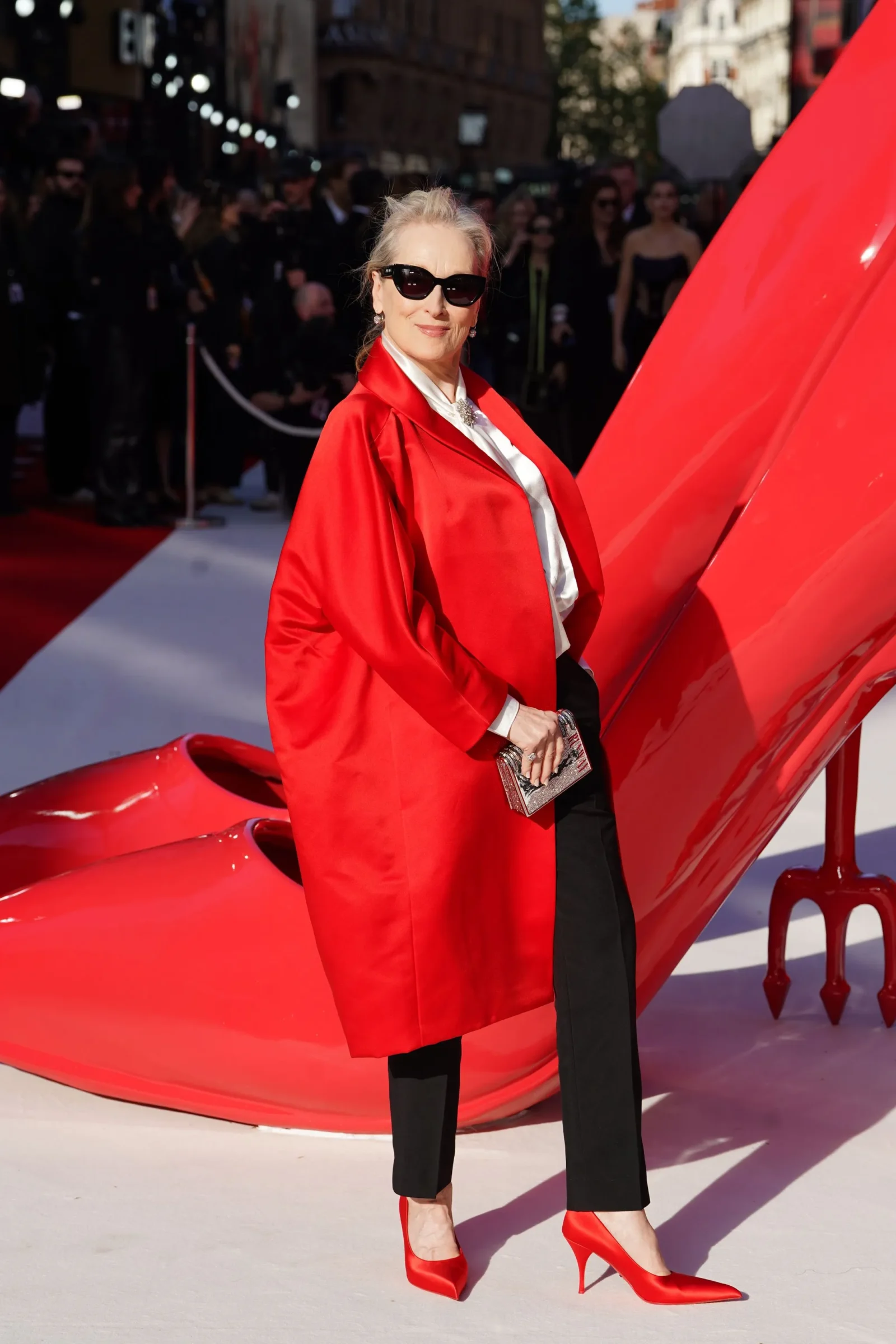 meryl streep in a bright red coat and red heels posing on a red carpet, sunglasses on, beside a large red sculpture on a sunny event day.