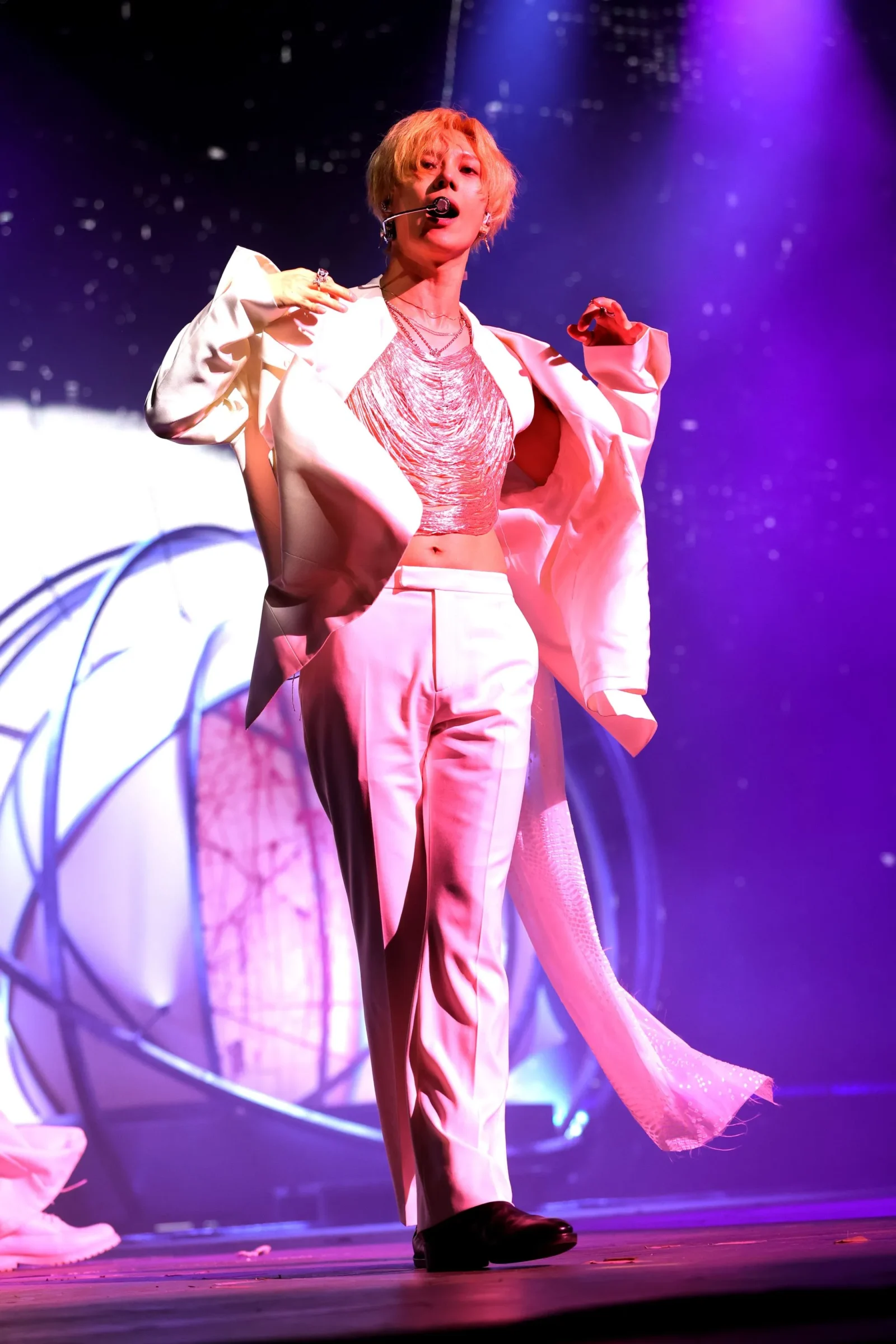 Performer on stage wearing a pink suit and metallic top, singing into a headset mic under purple spotlights.