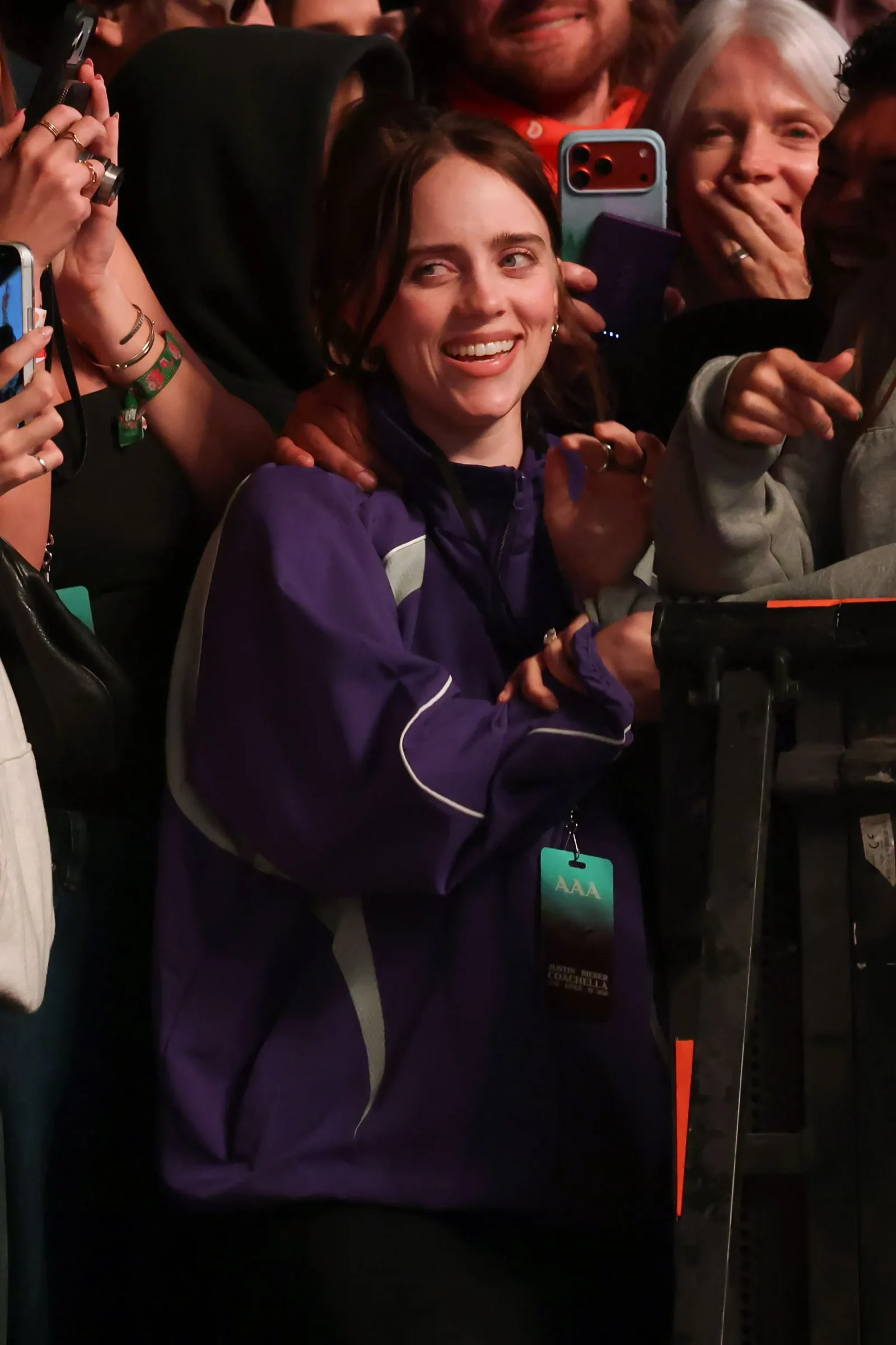 Smiling woman in a purple jacket among fans taking photos at a crowded event outside a barrier.